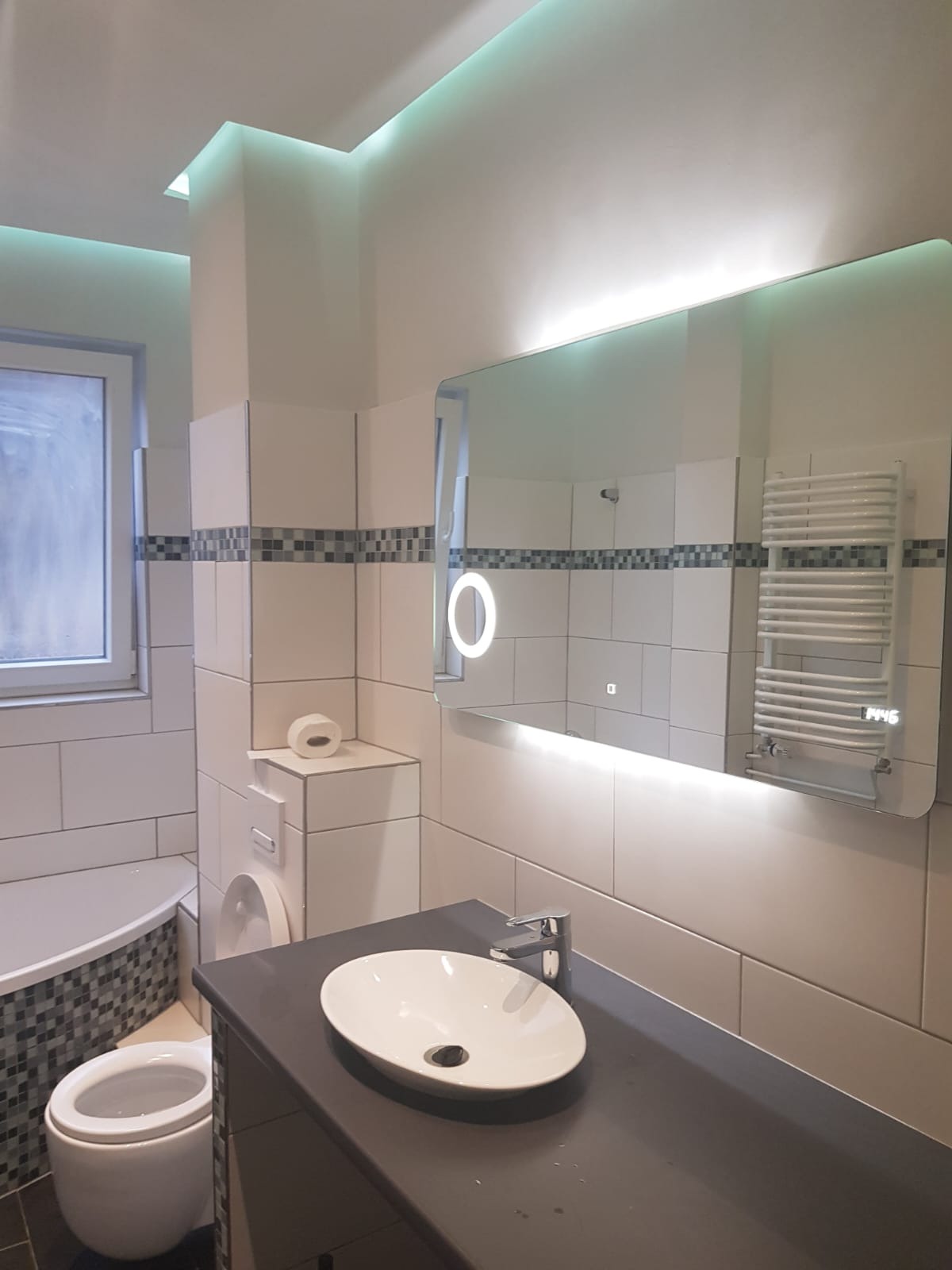 Nowoczesna łazienka z podświetlanym lustrem, grzejnikiem drabinkowym i umywalką nablatową na szarym blacie, wykończona białymi płytkami z dekoracyjnym pasem mozaiki.