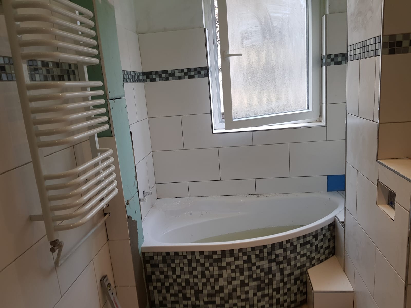 Łazienka w trakcie remontu: białe płytki ścienne z dekoracyjnym pasem mozaiki, obudowana mozaiką wanna wypełniona wodą, grzejnik drabinkowy, widoczne elementy instalacji przed zabudową.