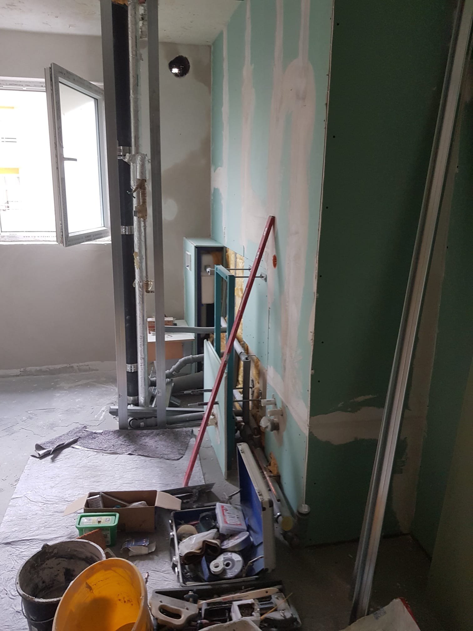 Wnętrze w trakcie prac remontowych: widoczne rury instalacyjne w metalowej ramie, fragment ściany z płyt gipsowo-kartonowych, narzędzia i materiały budowlane porozrzucane na podłodze przy oknie.