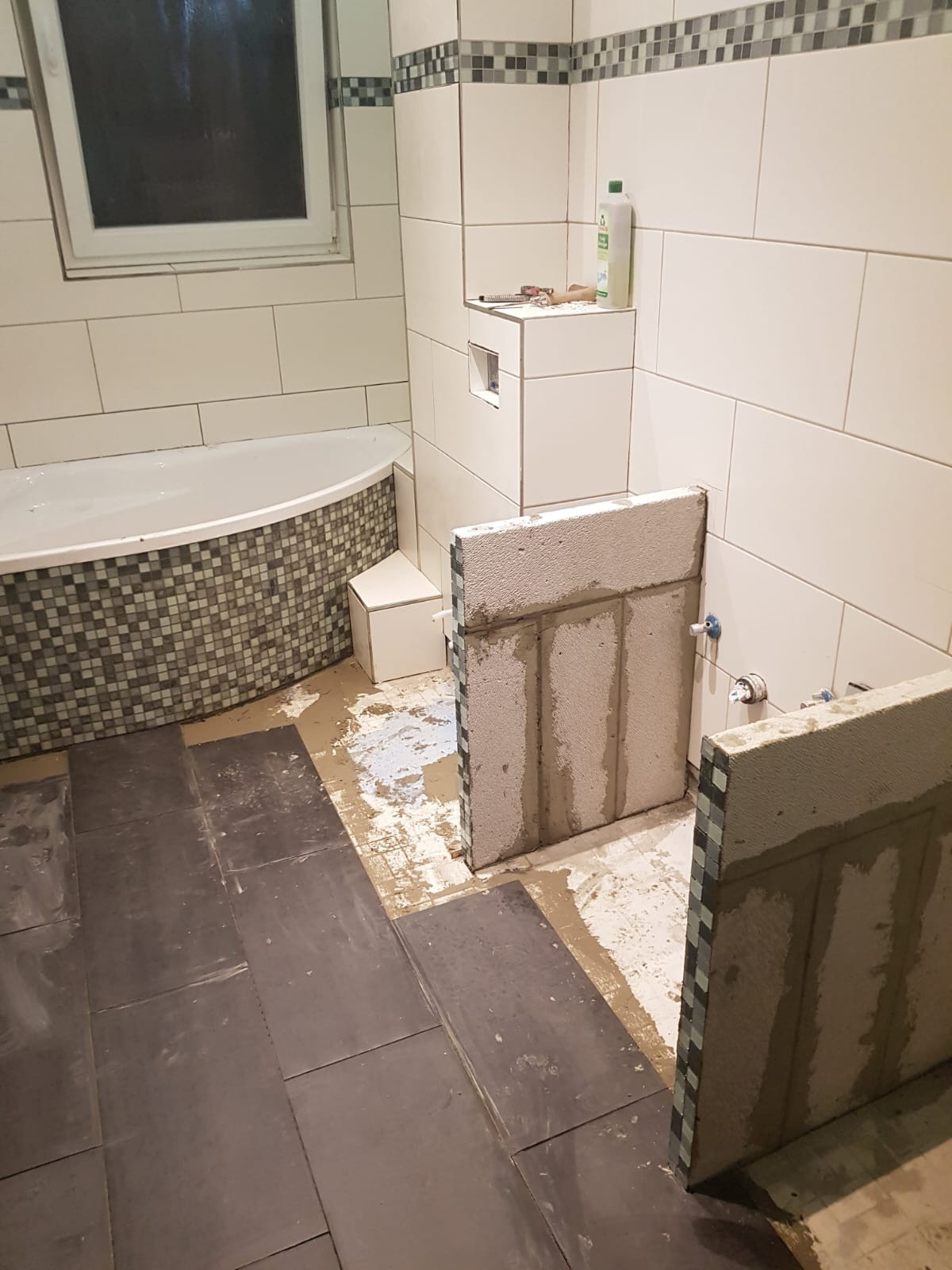 Łazienka w trakcie prac: układanie ciemnych płytek podłogowych, fragmentarycznie wzniesiona ścianka działowa z betonu komórkowego, widoczna wanna z mozaiką i świeżo położone płytki ścienne.