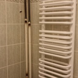 Ryszard Kościelny - Biały, drabinkowy grzejnik łazienkowy zamontowany na ścianie z zielono-beżowych płytek, z widocznymi rurami przyłączeniowymi z osadami.