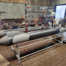 Ryszard Kościelny - Dwa metalowe, spawane wymienniki ciepła na białym stelażu w warsztacie. Widoczne rury, zawory, narzędzia i elementy instalacji. Surowe, przemysłowe otoczenie.