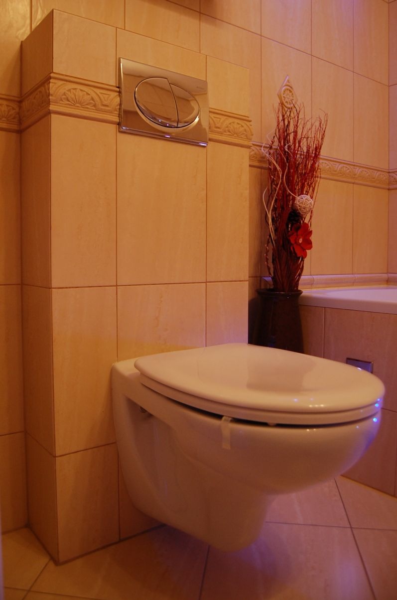 Wisząca toaleta z chromowanym przyciskiem spłukiwania na tle beżowych płytek, dekoracyjna kompozycja z suszonych gałązek i kwiatów w wazonie.