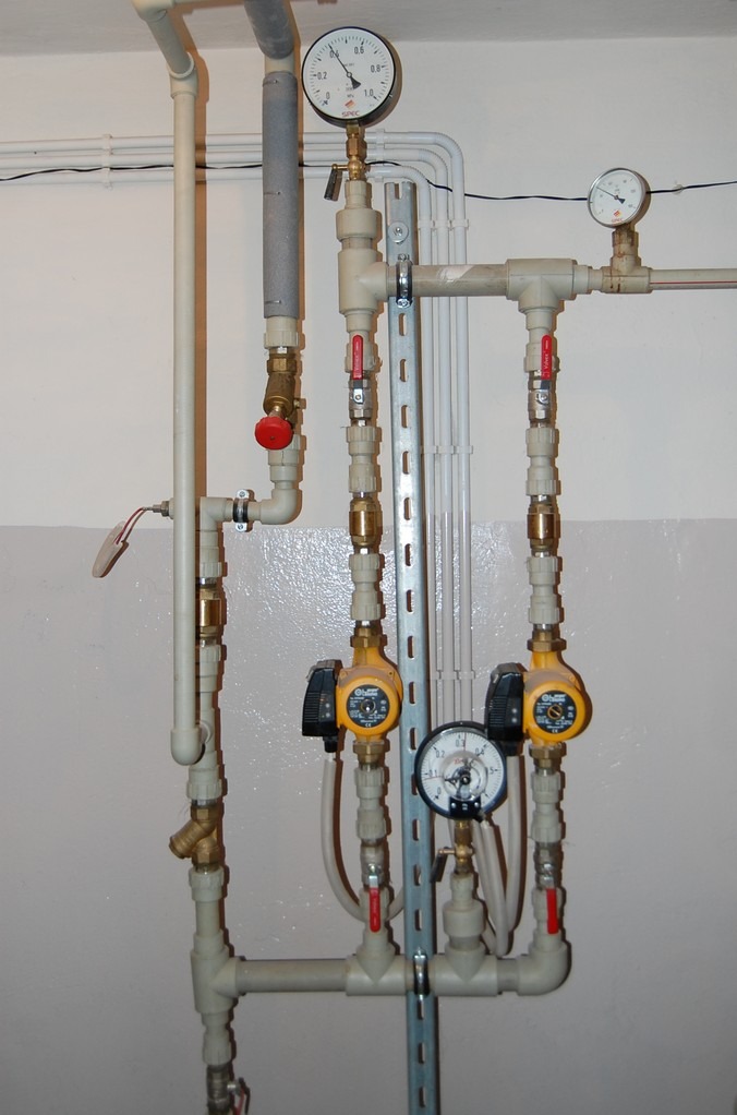 Kompleksowa instalacja z rur PCV, manometry i pompy obiegowe zamontowane na ścianie w piwnicy, widoczne liczne zawory kulowe z czerwonymi uchwytami.