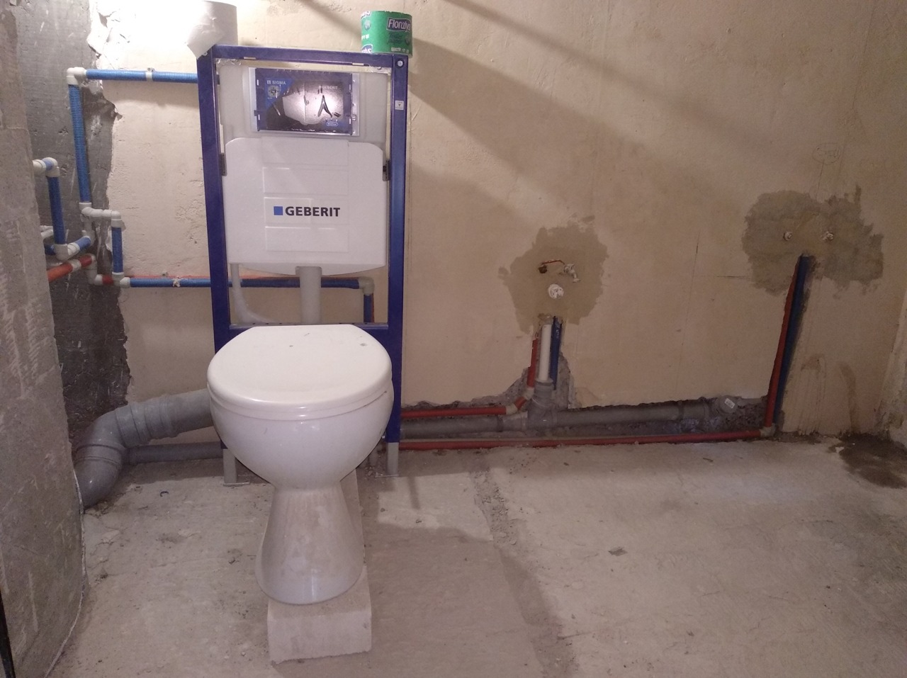 Biała toaleta kompaktowa marki Geberit, zamontowana na bloczku betonowym, z widocznymi instalacjami hydraulicznymi w surowym stanie, przed wykończeniem łazienki.