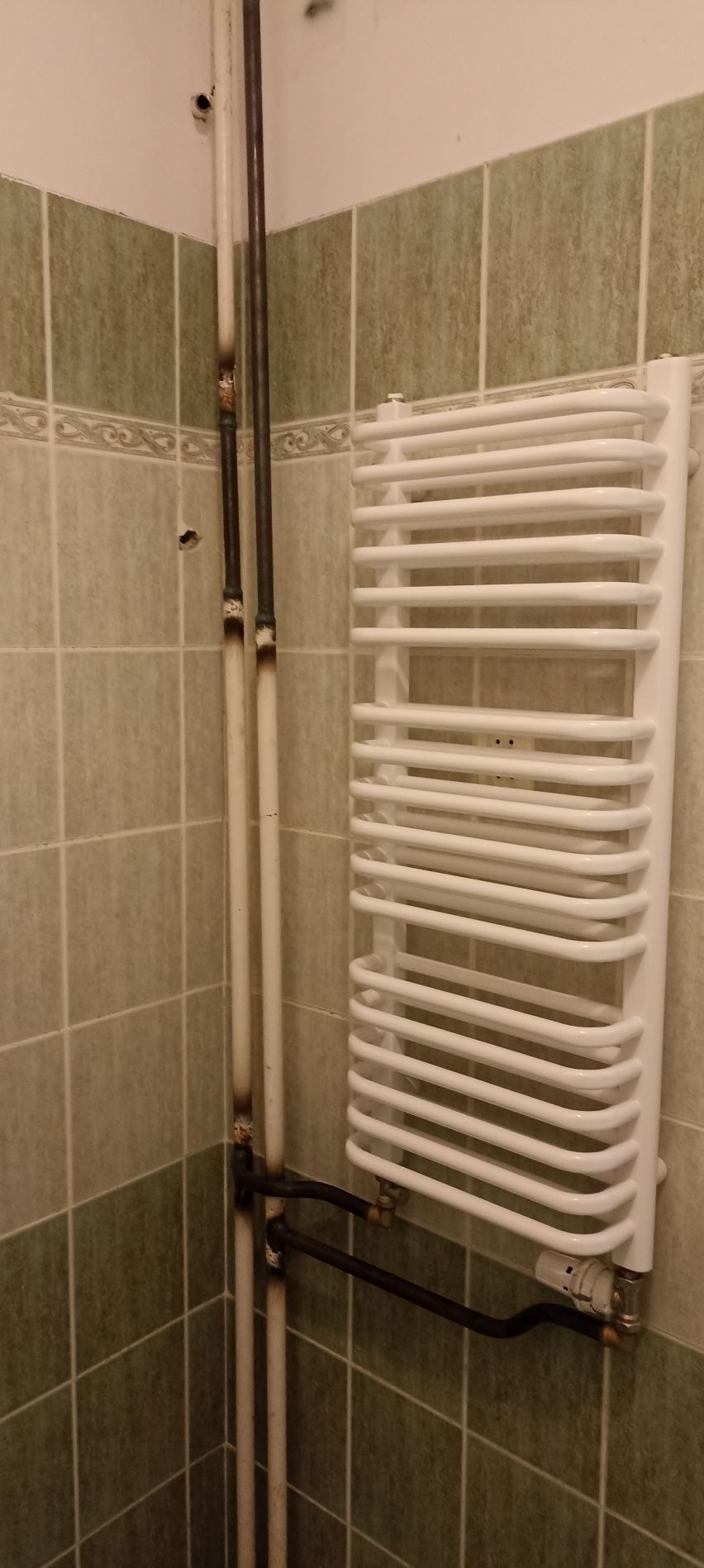 Biały, drabinkowy grzejnik łazienkowy zamontowany na ścianie z zielono-beżowych płytek, z widocznymi rurami przyłączeniowymi z osadami.
