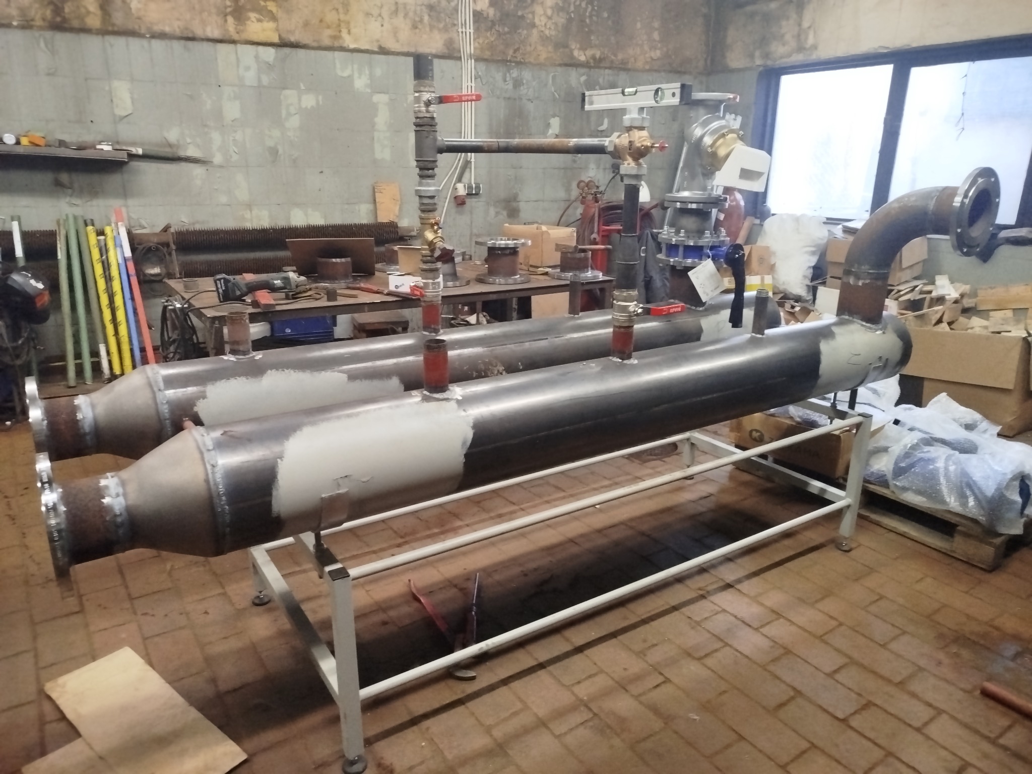 Dwa metalowe zbiorniki na białym stelażu w warsztacie, z widocznymi spawami i elementami instalacji. W tle narzędzia i okno, ceglana podłoga.