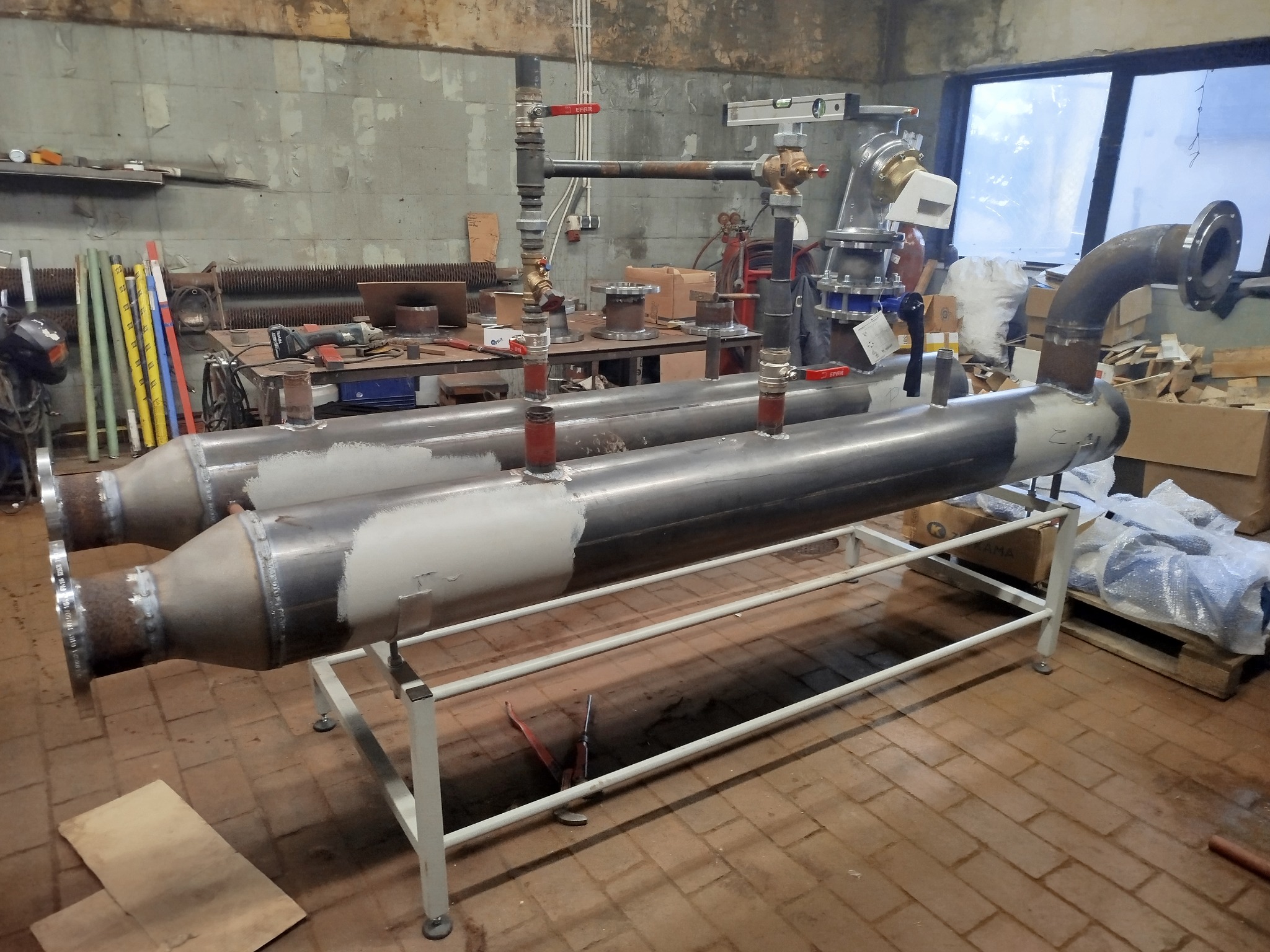 Dwa metalowe, spawane wymienniki ciepła na białym stelażu w warsztacie. Widoczne rury, zawory, narzędzia i elementy instalacji. Surowe, przemysłowe otoczenie.