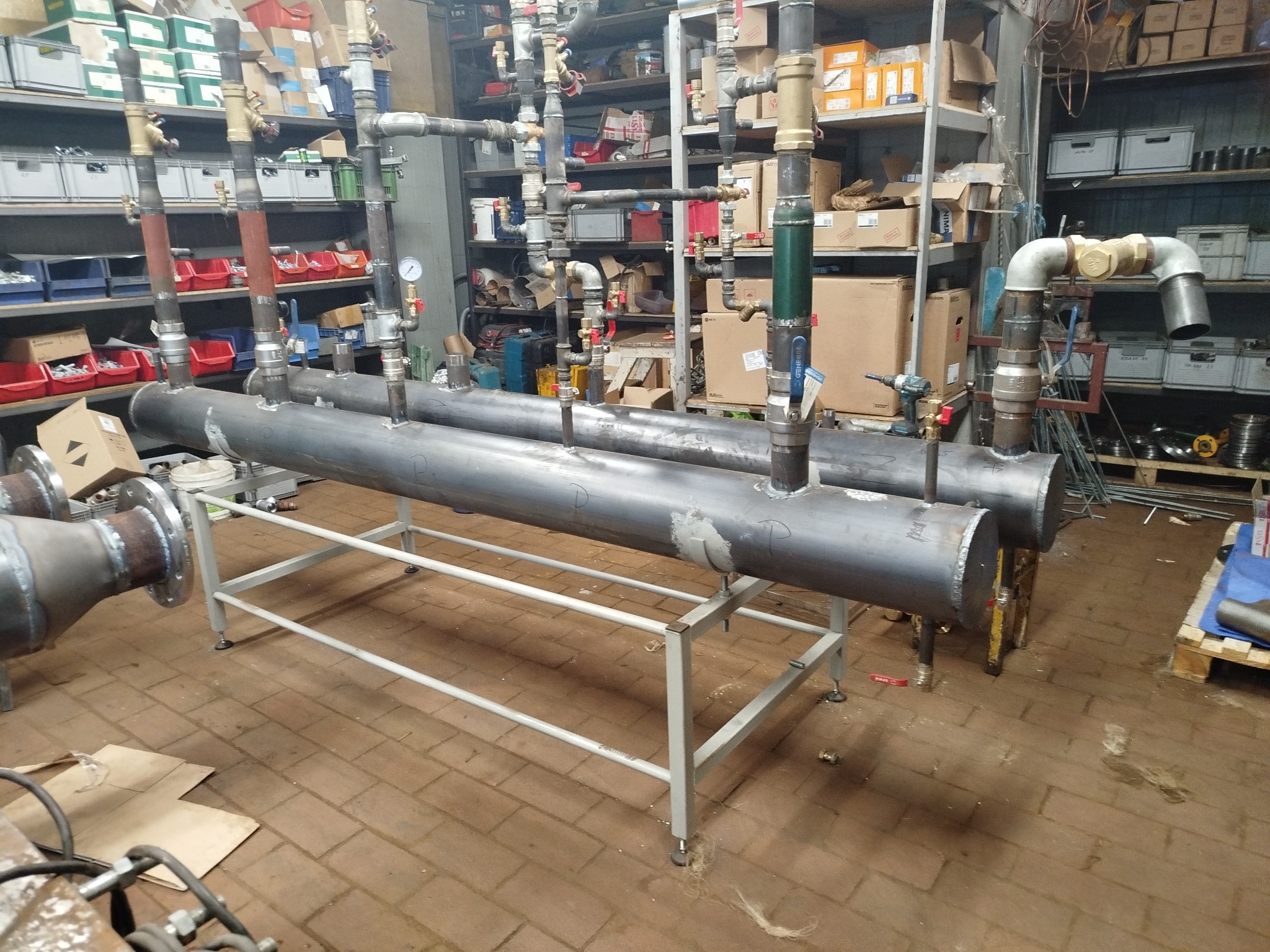 Dwa duże, metalowe zbiorniki na stelażu z podłączonymi rurami i zaworami, w tle regały z narzędziami i materiałami. Widoczne ślady spawania na zbiornikach.