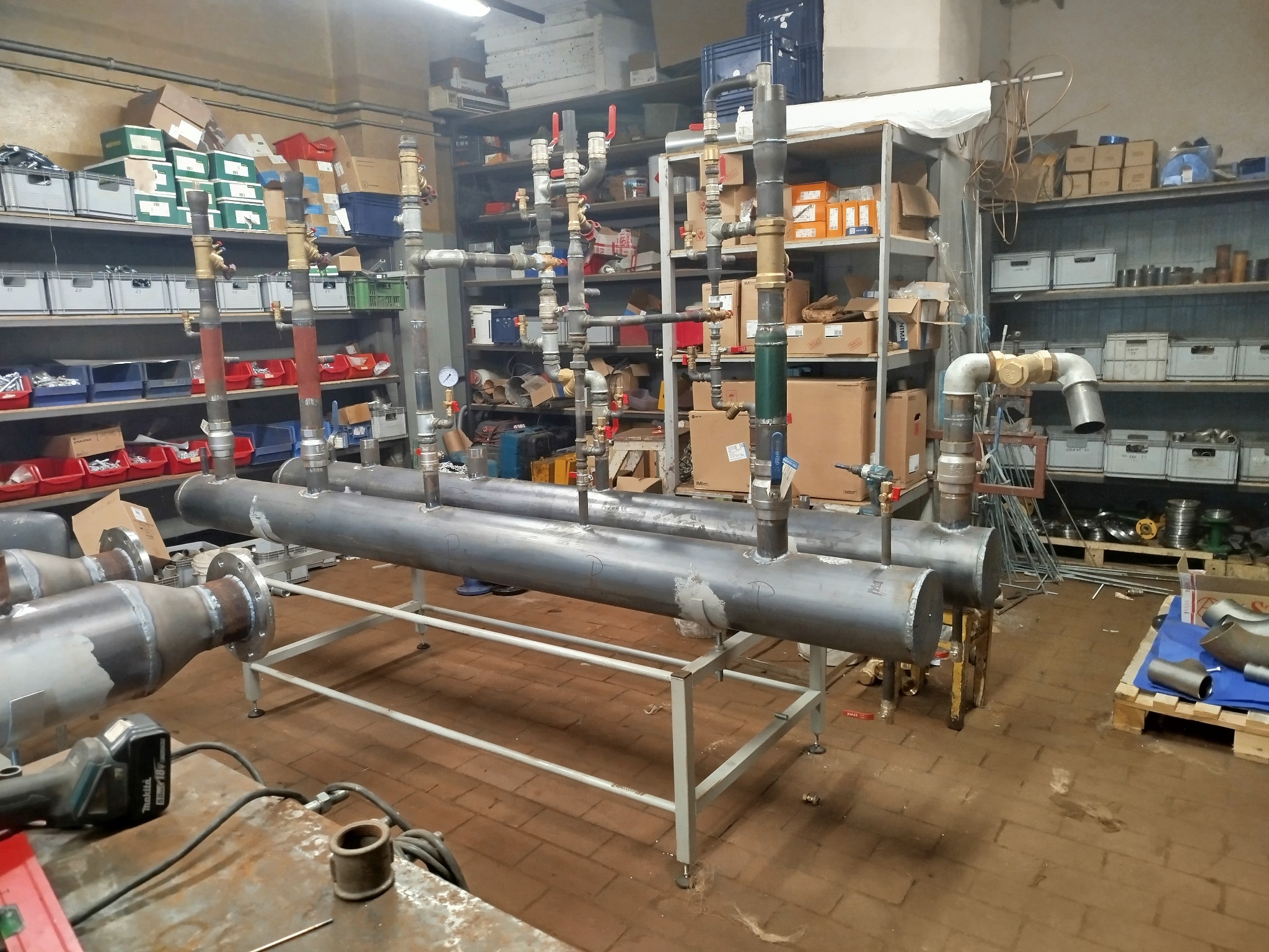 Warsztatowa instalacja grzewcza: Dwa poziome, szare zbiorniki na metalowym stelażu, z licznymi pionowymi rurami i zaworami. W tle regały z narzędziami i materiałami.