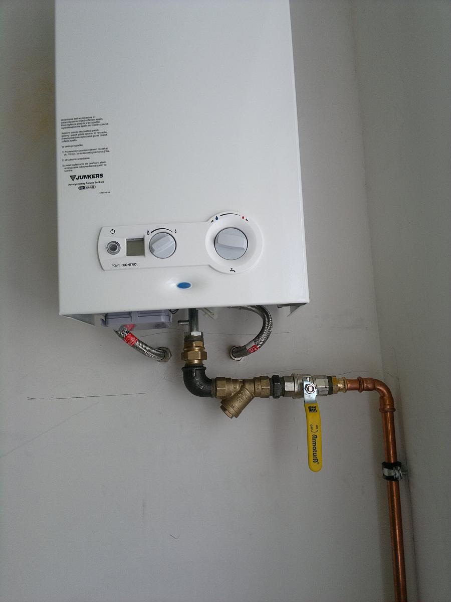 Biały piec gazowy Junkers zamontowany na ścianie, z widocznymi przyłączami hydraulicznymi i zaworem odcinającym. Instalacja w jasnym pomieszczeniu.