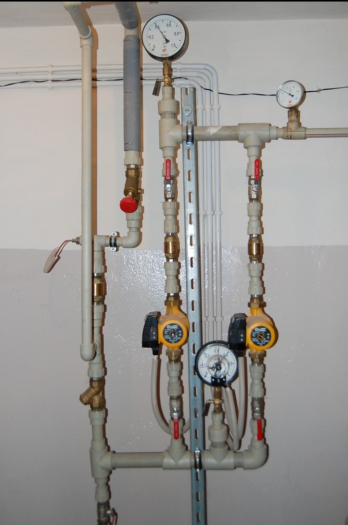 Instalacja CO z dwoma pompami obiegowymi, manometrami i zaworami, zamontowana na ścianie. Widoczne rury z tworzywa i metalowe elementy mocujące.