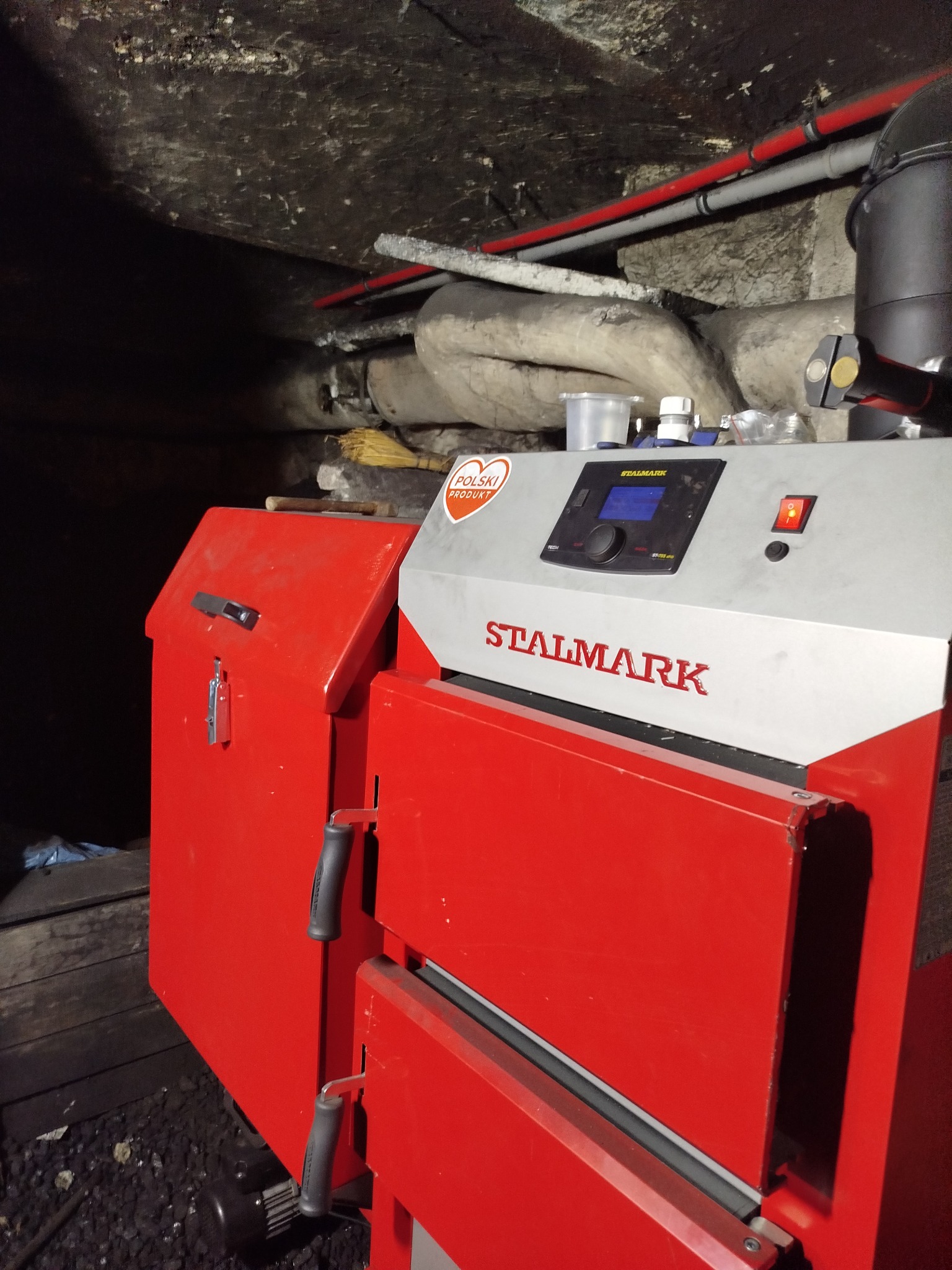 Czerwono-szary piec marki Stalmark w kotłowni. Widoczne rury instalacji grzewczej i elementy sterujące piecem. Oświetlenie pomieszczenia jest słabe.