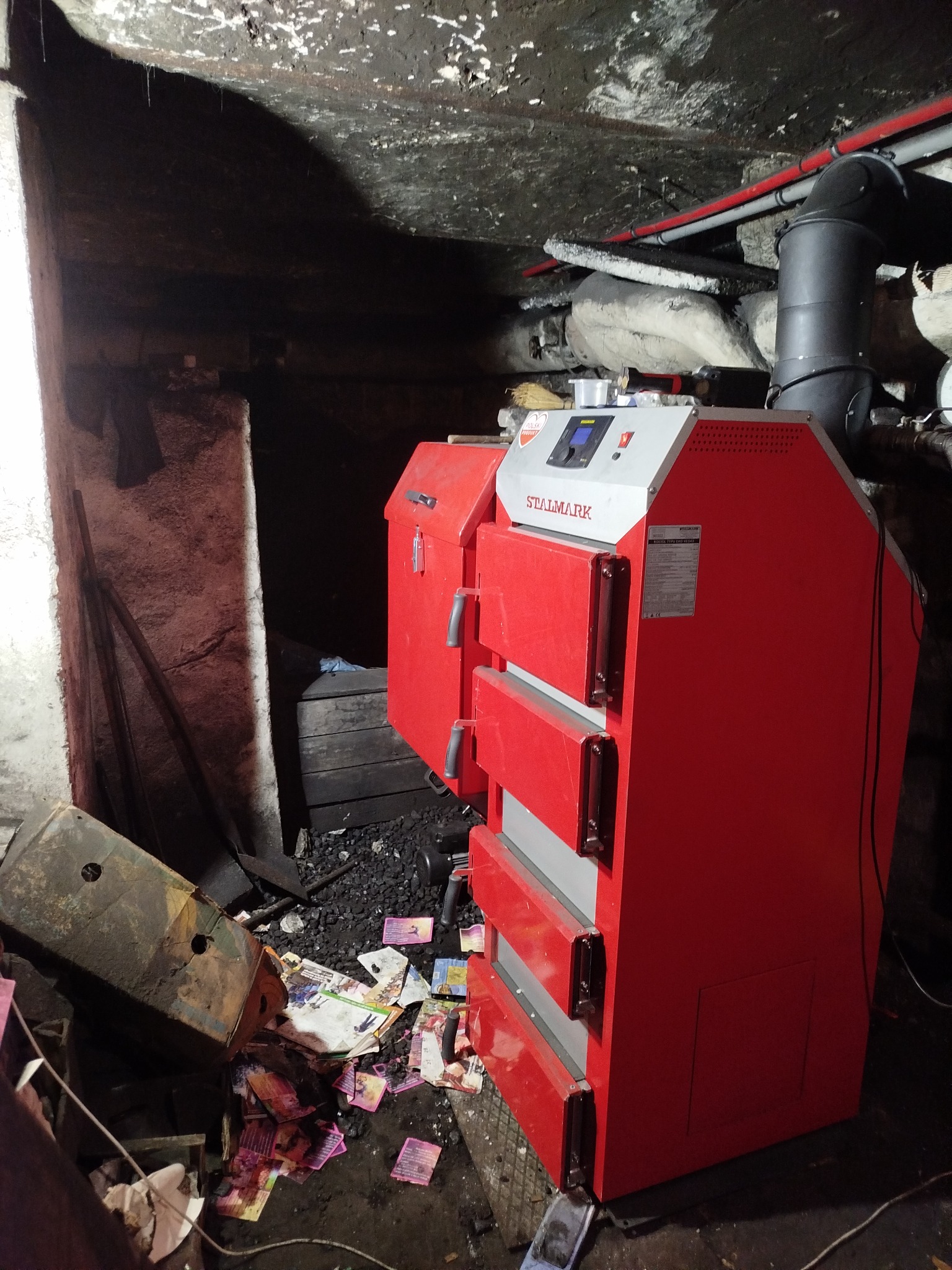 Czerwony piec Stalmark w piwnicy, widoczne rury instalacji grzewczej i węgiel. Nierówne oświetlenie, surowy klimat kotłowni.