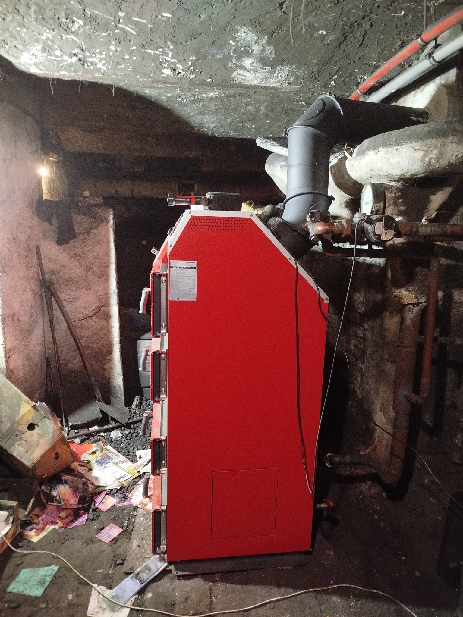 Czerwony piec CO z podajnikiem, zamontowany w piwnicy w Mikołowie. Widoczne rury instalacji grzewczej i węgiel. Nowoczesny piec w surowym otoczeniu.