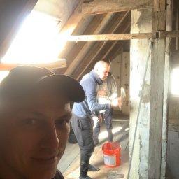 Majster Wrocław - Mężczyzna robiący selfie na poddaszu podczas prac remontowych, w tle drugi mężczyzna skuwa tynk młotkiem, widoczne odsłonięte drewniane elementy konstrukcyjne dachu, wiadro z zaprawą i narzędzia.
