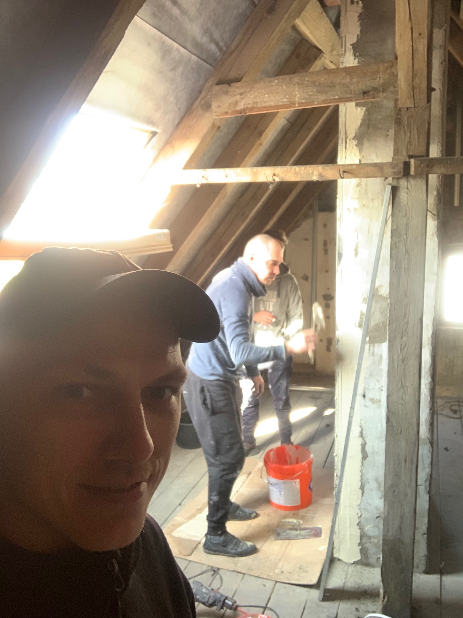 Mężczyzna robiący selfie na poddaszu podczas prac remontowych, w tle drugi mężczyzna skuwa tynk młotkiem, widoczne odsłonięte drewniane elementy konstrukcyjne dachu, wiadro z zaprawą i narzędzia.