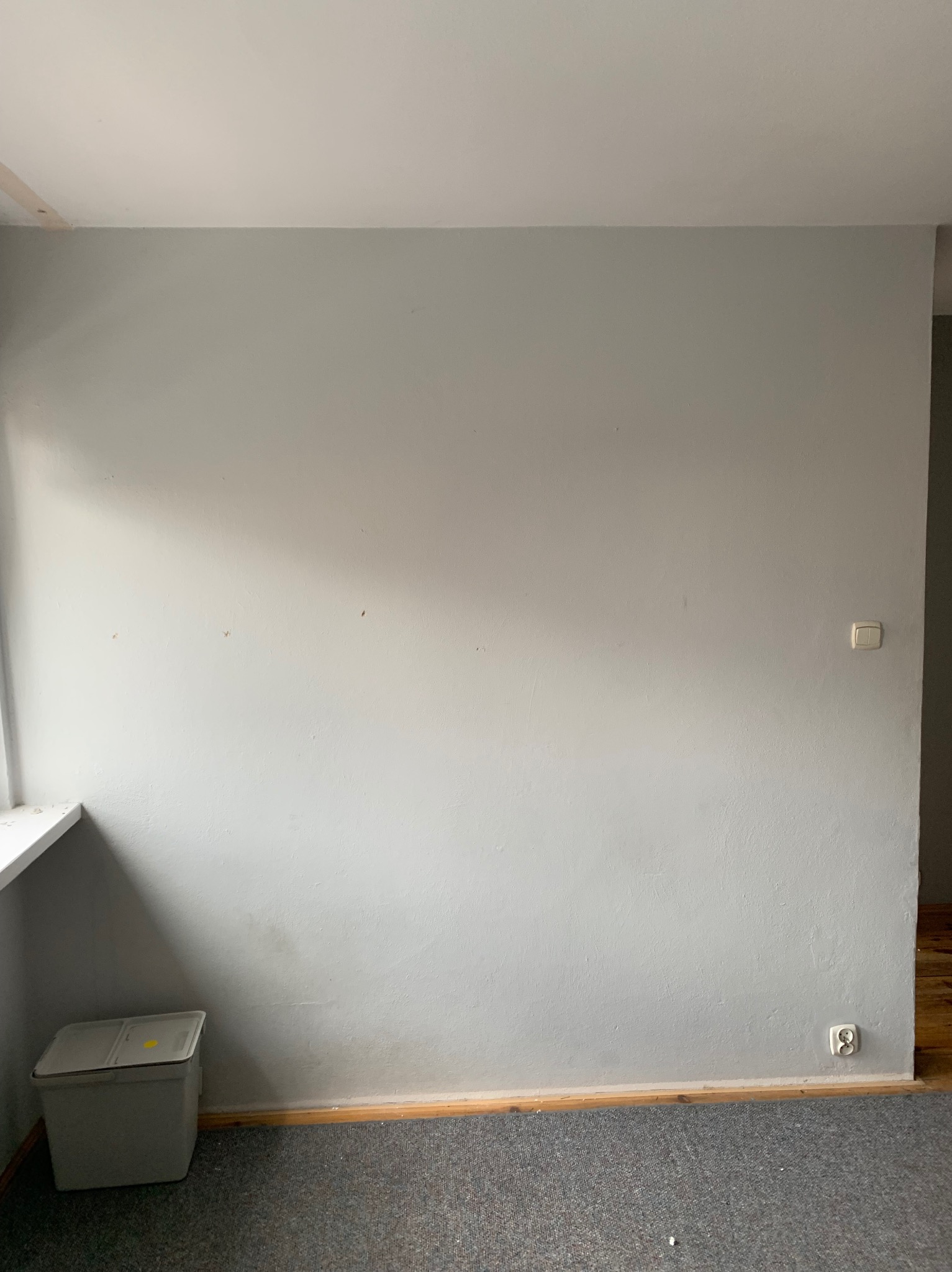 Szara ściana w trakcie przygotowania do remontu, widoczne ślady po usuniętych elementach, listwa przypodłogowa, gniazdko elektryczne i włącznik światła, na podłodze szara wykładzina, w rogu...