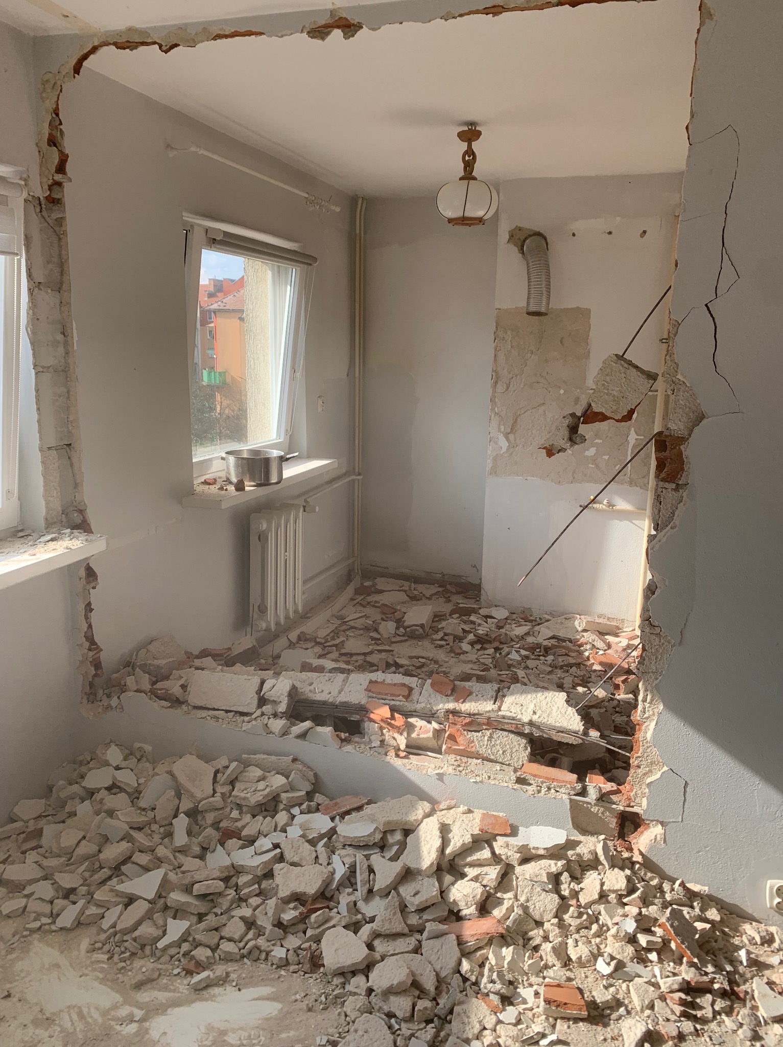 Wnętrze po wyburzeniu ściany działowej, widoczne gruzowisko, odsłonięte fragmenty instalacji, okno z garnkiem na parapecie, wisząca lampa i fragment komina wentylacyjnego.