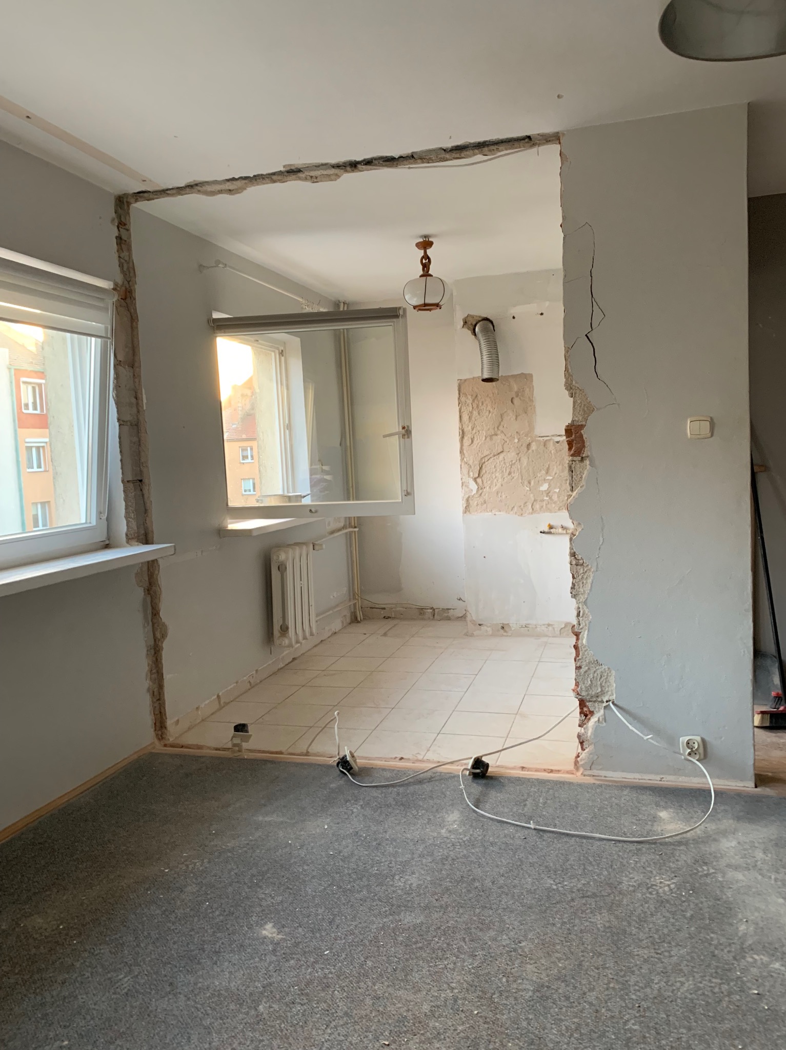 Wnętrze mieszkania w trakcie remontu, z widocznym otworem po wyburzonej ścianie, odsłonięte cegły i instalacje elektryczne, widok na sąsiednie pomieszczenie z oknem i grzejnikiem, na podłodze szary...