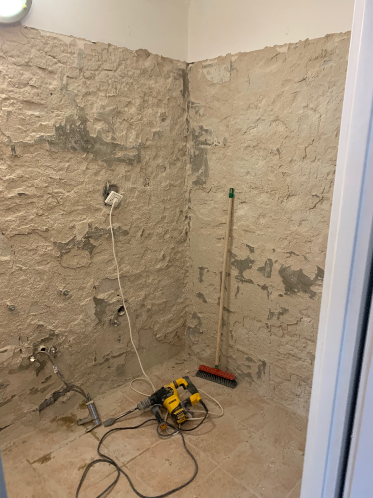 Wnętrze pomieszczenia w trakcie prac remontowych, ściany częściowo obdarte z tynku, widoczny kabel zasilający, wiertarka udarowa oraz zmiotka oparta o ścianę.