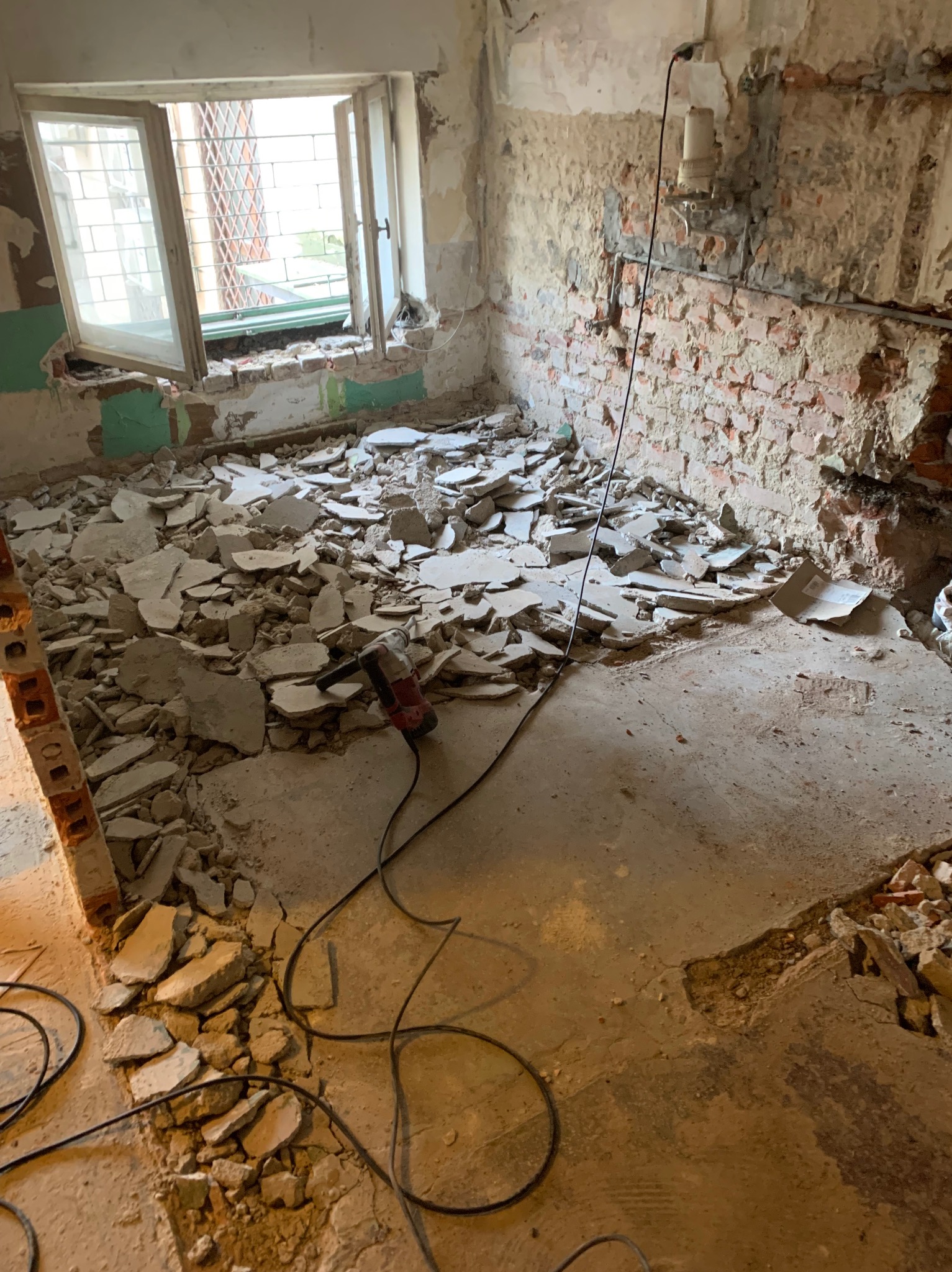 Wnętrze w trakcie prac rozbiórkowych; widoczne gruz, odsłonięte cegły, fragment okna z kratą i narzędzie do wyburzania. Oświetlenie naturalne wpada przez okno.