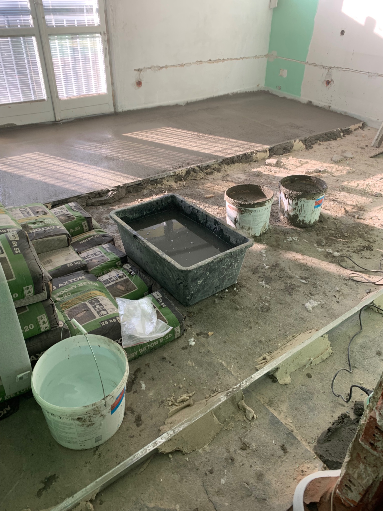 Wylewka betonowa na podłodze w trakcie realizacji, widoczne worki z zaprawą, pojemniki z wodą i zacierkami, okno z żaluzjami w tle.