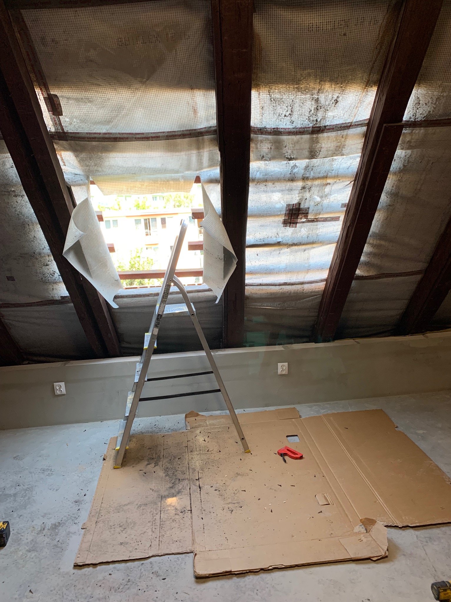Wnętrze poddasza z drewnianą konstrukcją dachu, zabezpieczoną folią, częściowo zerwaną, z widoczną drabiną aluminiową i kartonami na podłodze, przygotowane do prac remontowych.