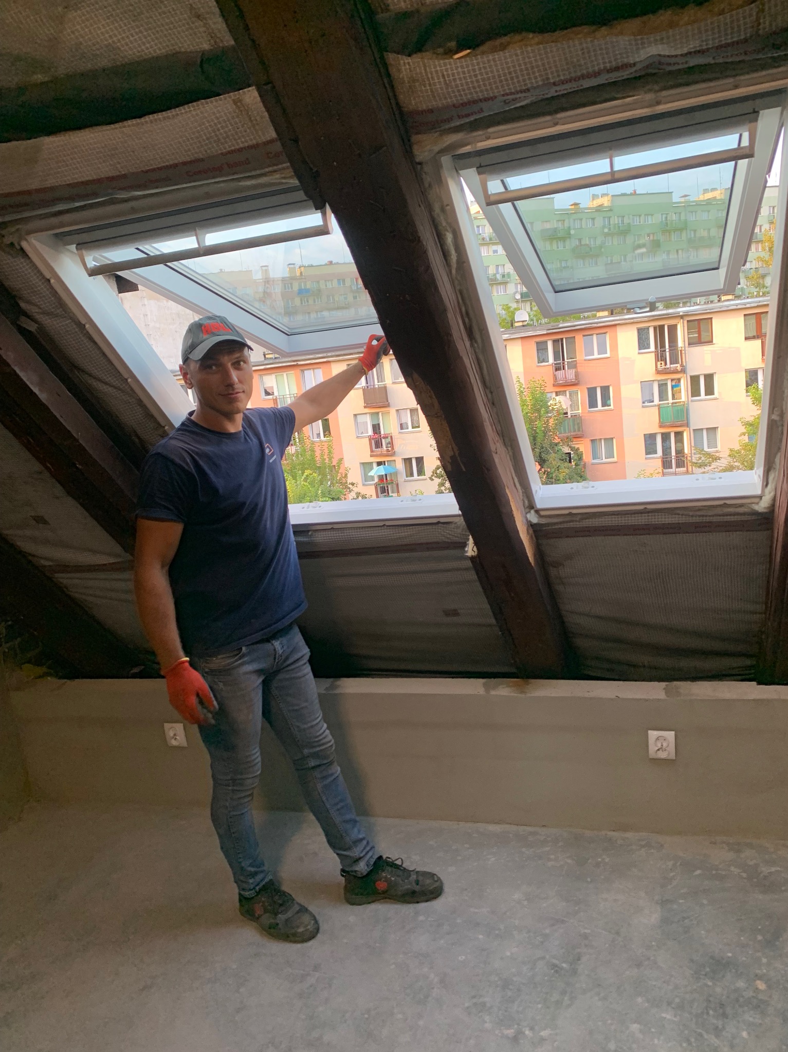 Mężczyzna w roboczych rękawicach prezentuje nowo zamontowane okno dachowe w surowym wnętrzu poddasza, z widokiem na budynki mieszkalne na zewnątrz.
