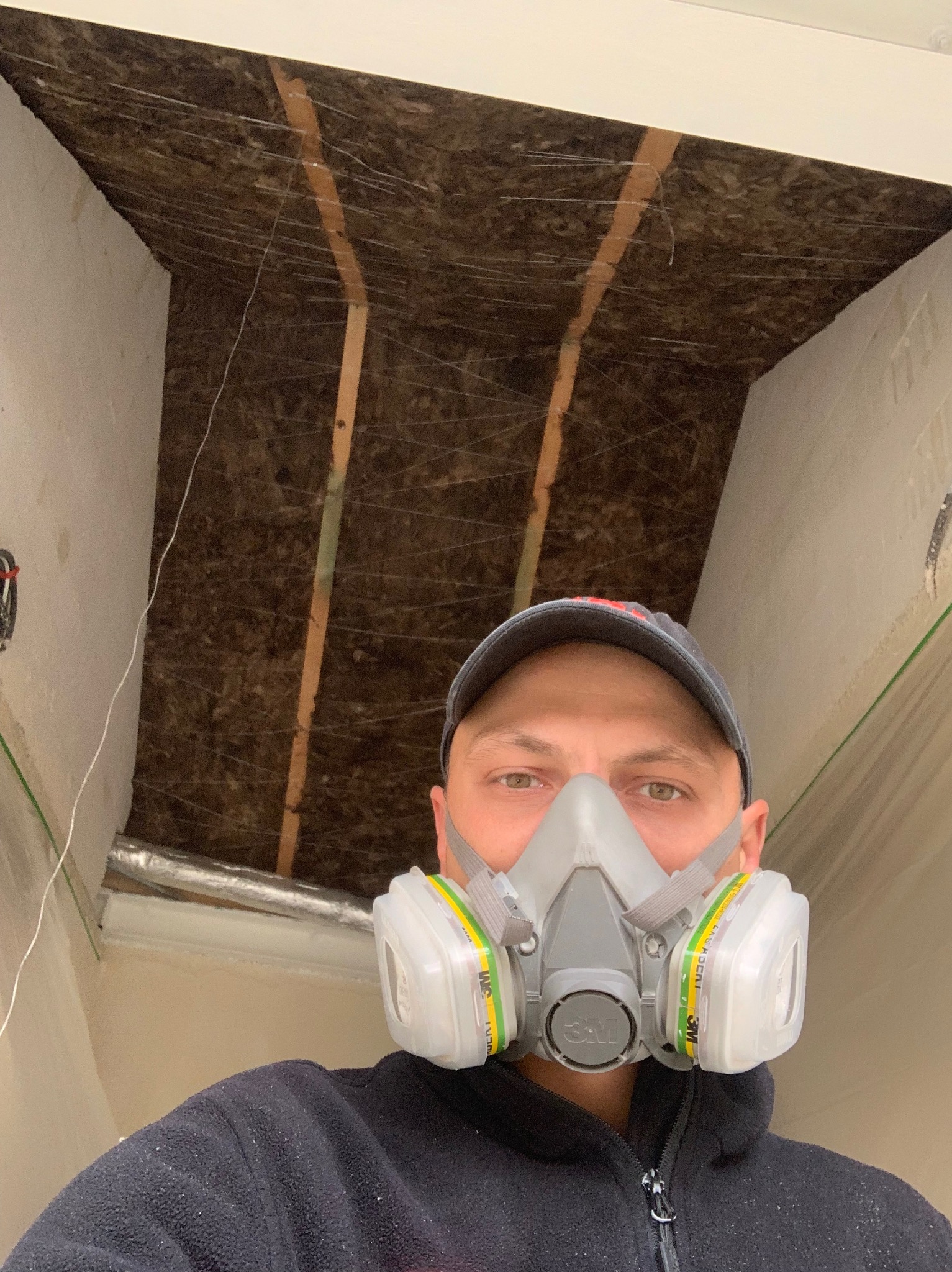 Mężczyzna w masce ochronnej 3M, patrzący w obiektyw, z widoczną izolacją z wełny mineralnej na suficie i fragmentem świeżo pomalowanej ściany.