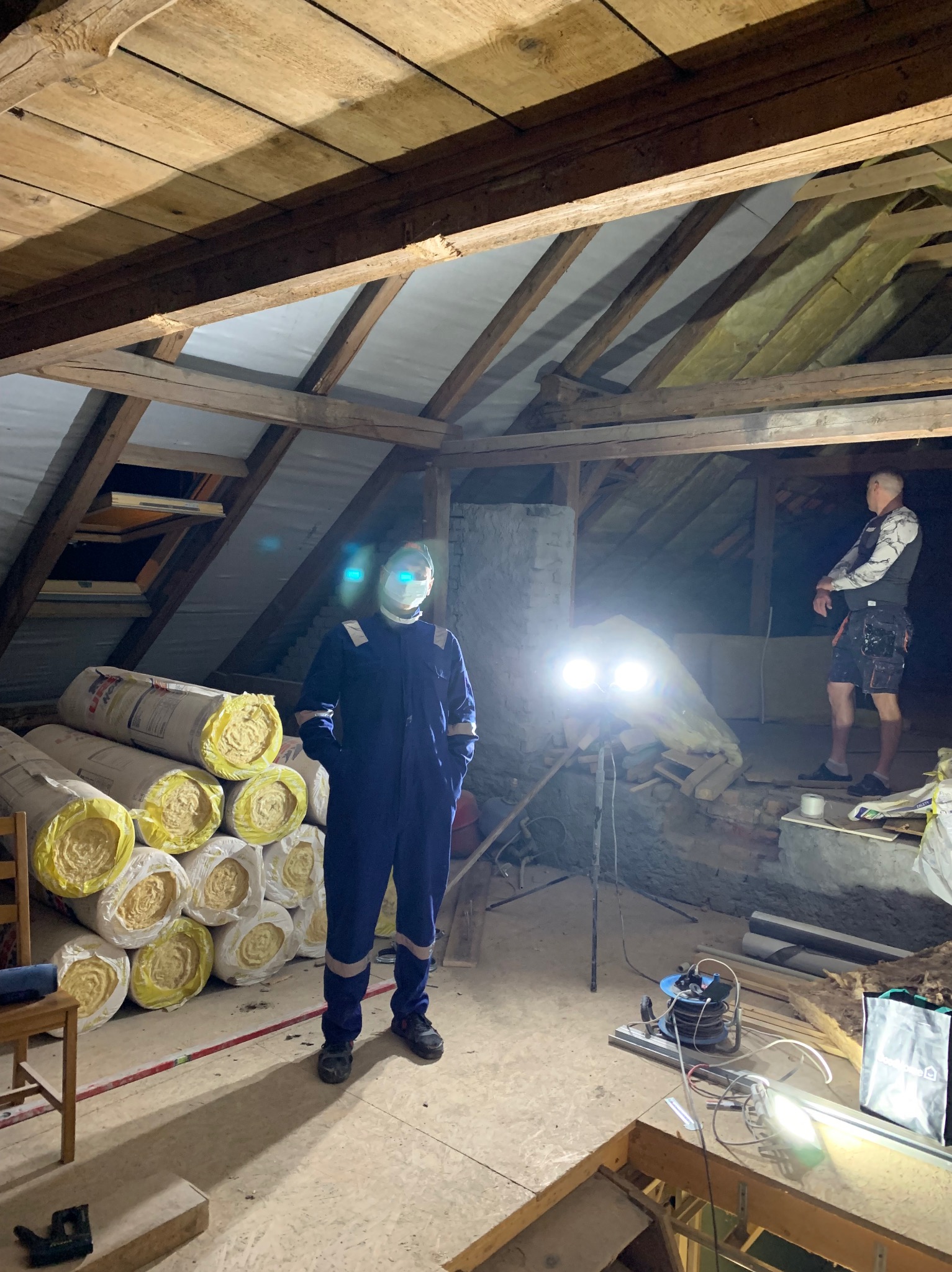 Poddasze w trakcie prac remontowych: pracownik w kombinezonie i masce stoi obok izolacji z wełny mineralnej, widoczne drewniane elementy konstrukcji dachu i prowizoryczne oświetlenie.