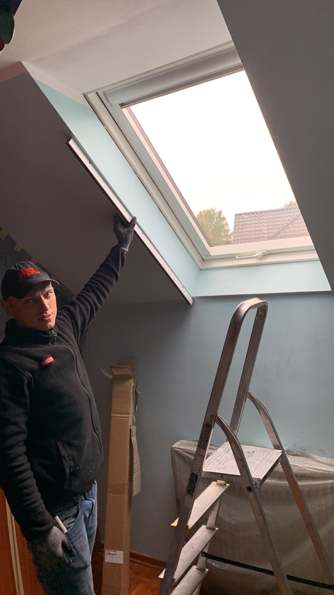 Monter w czarnej bluzie i czapce z daszkiem, z podniesioną ręką w rękawicy, instaluje okno dachowe; w tle drabina i materiały budowlane.