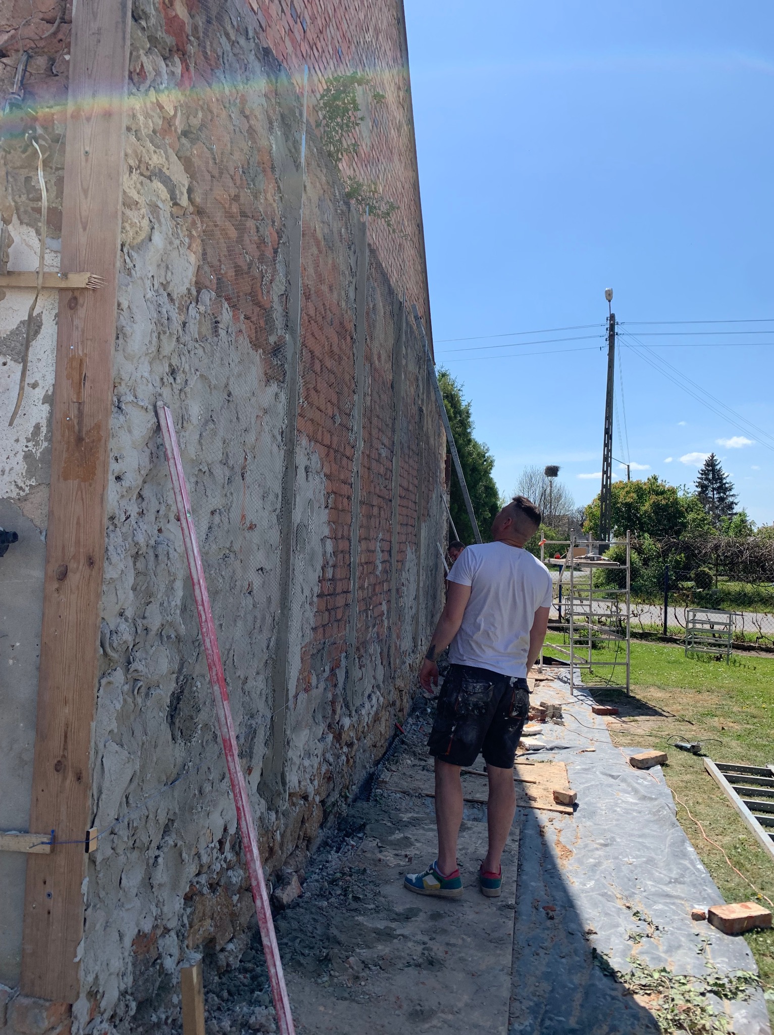 Mężczyzna w białej koszulce i wzorzystych spodenkach, z profilu, pracuje przy zewnętrznej ścianie budynku z odsłoniętą cegłą i fragmentami starego tynku, zabezpieczonej siatką; widoczne rusztowania...