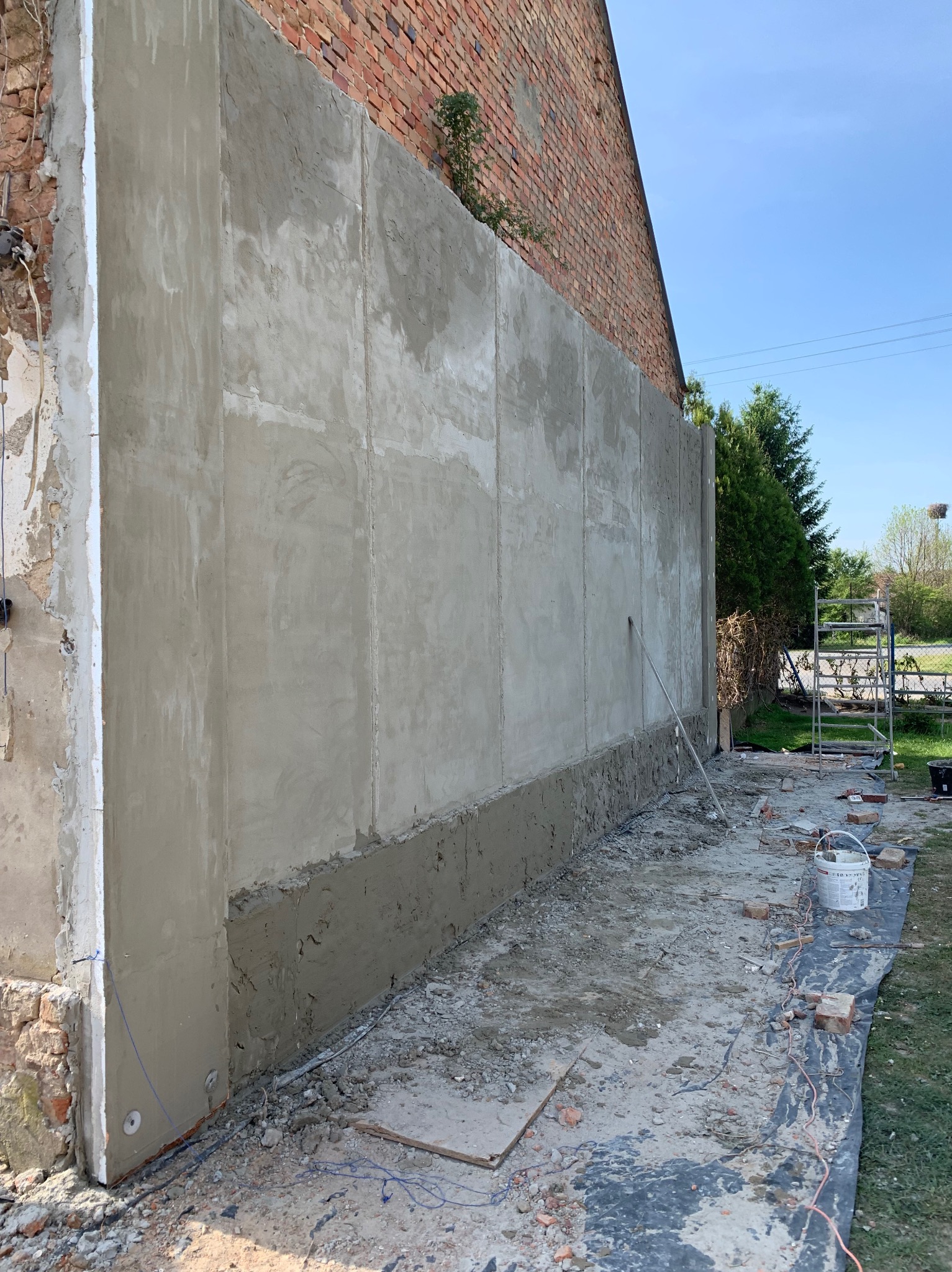 Świeżo otynkowana ściana budynku z widocznymi śladami prac murarskich, rusztowaniem i narzędziami na zewnątrz, częściowo odsłonięta cegła na sąsiedniej ścianie, słoneczny dzień.