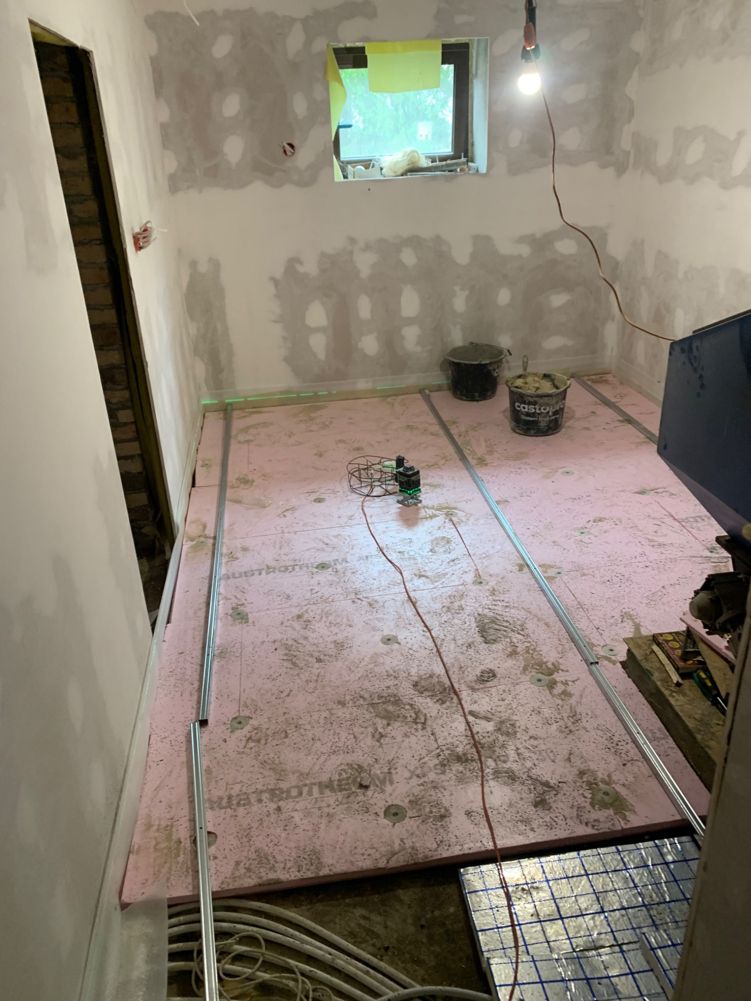 Przygotowanie do wylewki podłogi: różowy styropian, profile, poziomica laserowa. Ściany w trakcie szpachlowania, widoczne kable instalacji.