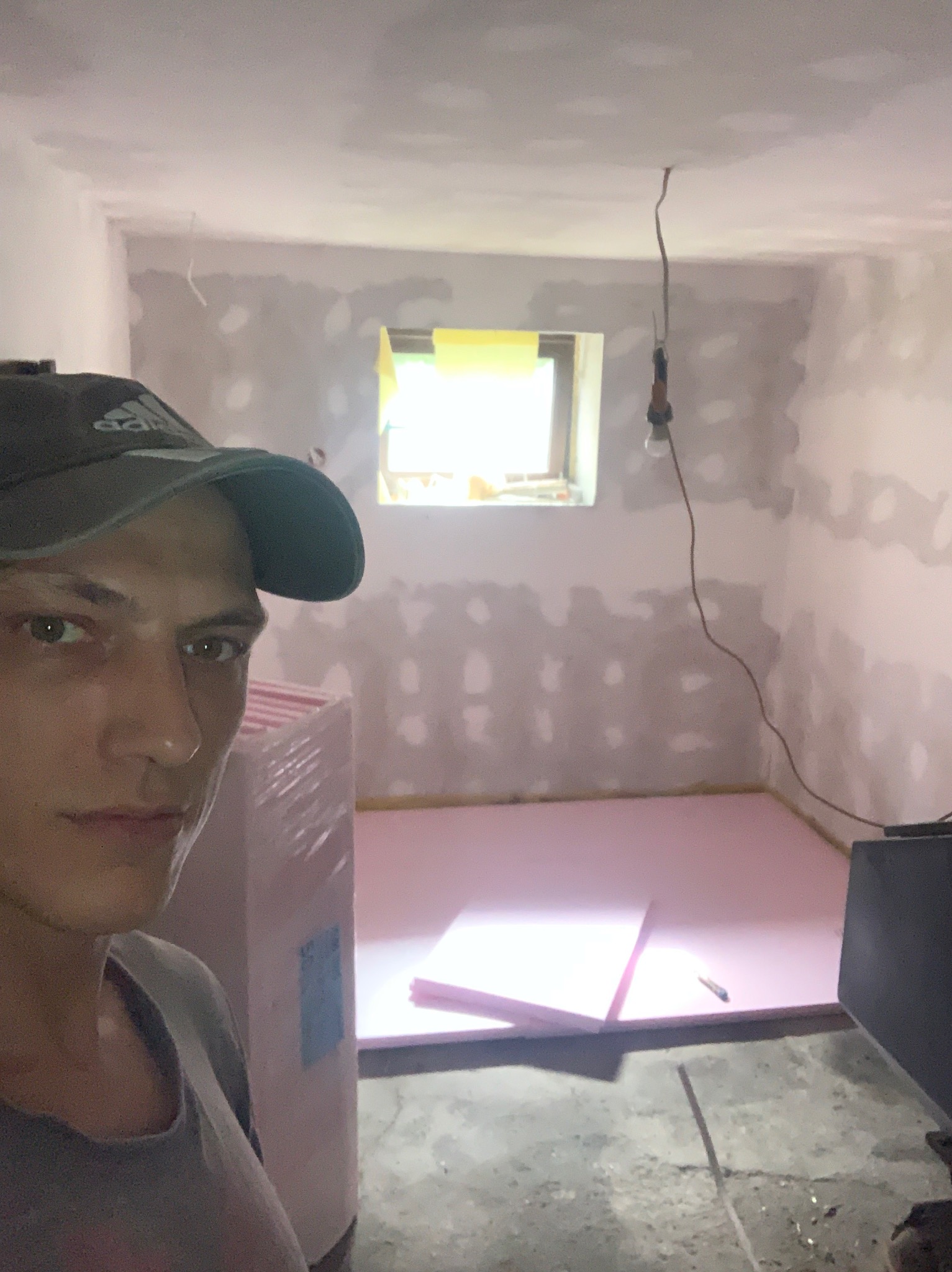 Układanie różowego styropianu na podłodze remontowanego pokoju z oknem. Widoczna osoba w czapce z daszkiem i surowy stan ścian z widocznymi łatami gipsowymi.