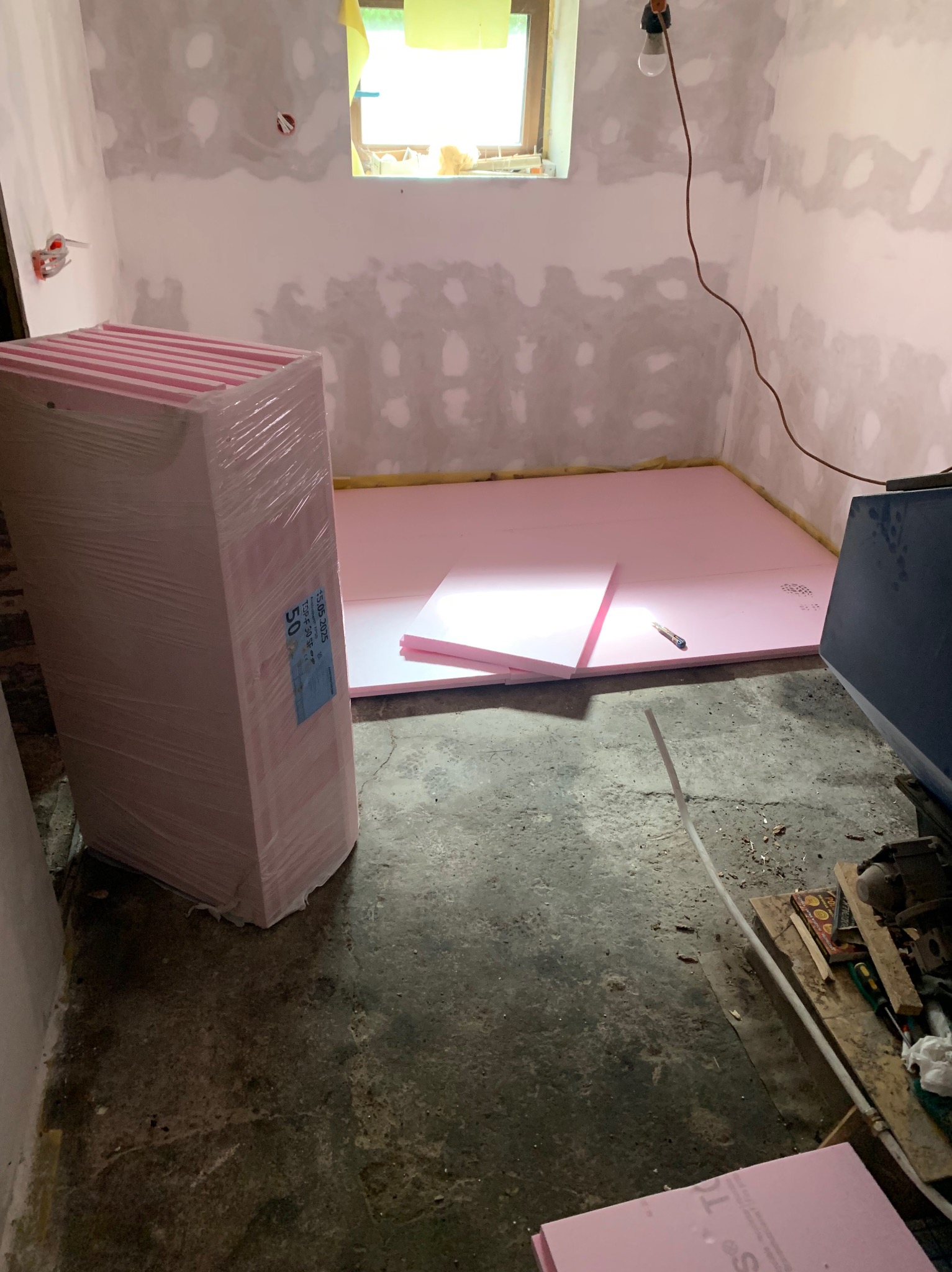 Przygotowanie do izolacji podłogi: różowe płyty styropianowe rozłożone w surowym pomieszczeniu z oknem, obok zapakowany stos płyt i nóż do cięcia.