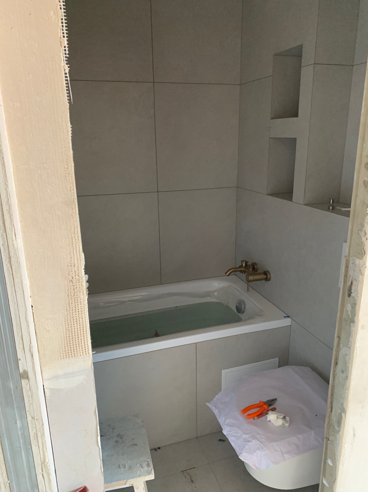 Remont łazienki: wykończona wanna z baterią w kolorze złota, obłożona jasnymi płytkami. Widoczne narzędzia i fragment toalety zabezpieczonej folią.