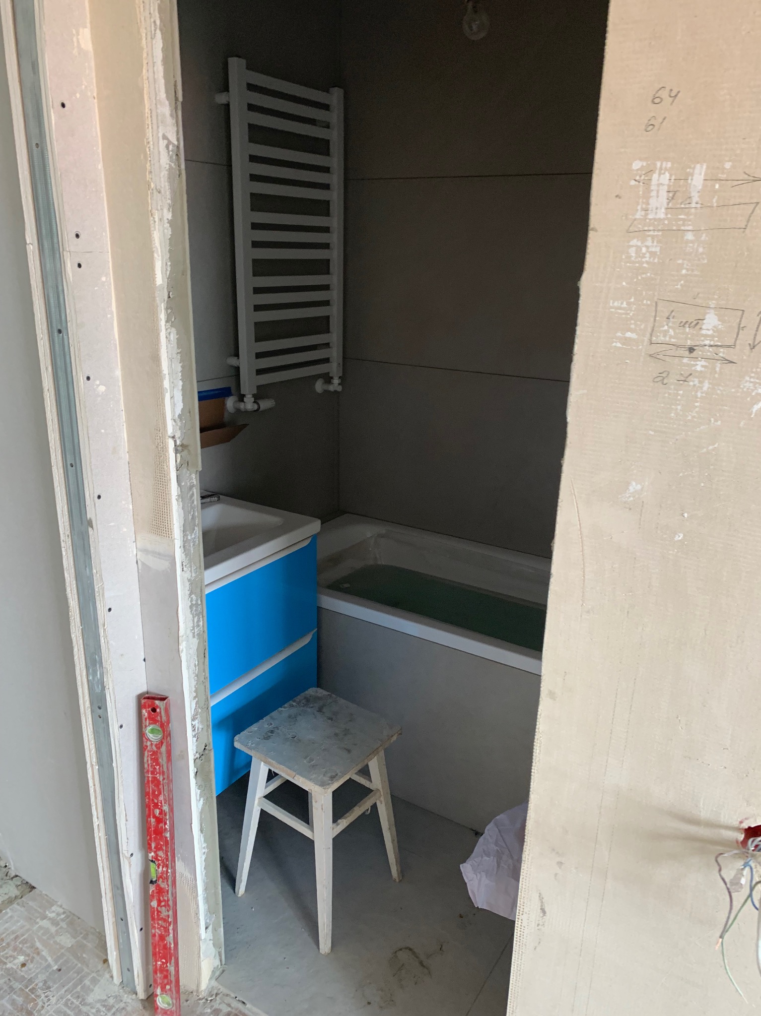 Niedokończona łazienka z niebieską szafką pod umywalkę, drabinkowym grzejnikiem i taboretem. Widoczne elementy instalacji i poziomica.