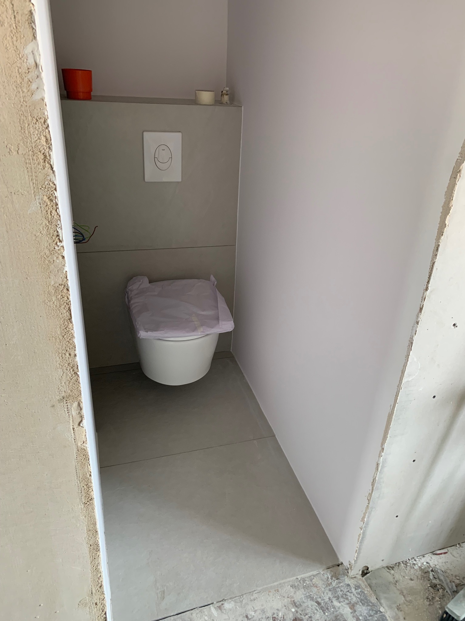 Nowoczesna toaleta w trakcie remontu, z zakrytym sedesem i widocznymi elementami instalacji. Szare płytki i surowy stan ścian.