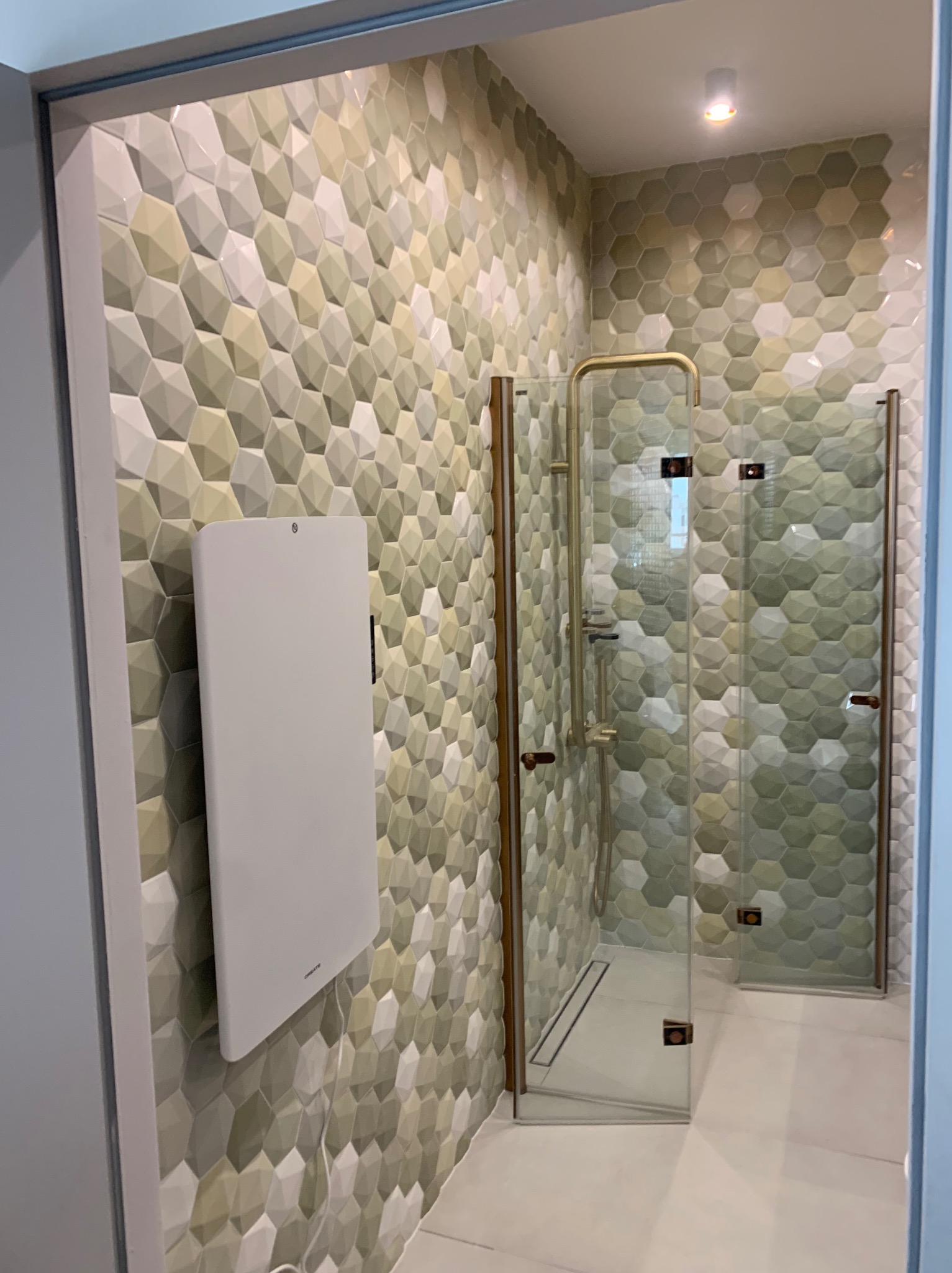 Nowoczesna kabina prysznicowa ze złotymi detalami i heksagonalnymi płytkami w odcieniach zieleni i bieli. Na ścianie zamontowany biały grzejnik.
