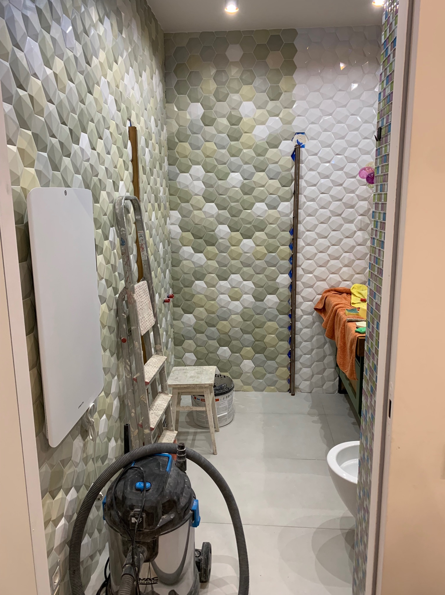 Łazienka w trakcie remontu: heksagonalne płytki 3D na ścianach, drabina, grzejnik, odkurzacz budowlany. Widok zza drzwi.