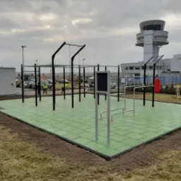 Zewnętrzna siłownia z zieloną nawierzchnią z gumowych płyt, widoczne stalowe konstrukcje do ćwiczeń, w tle budynek lotniska z wieżą kontroli lotów i pracownicy w odblaskowych kamizelkach.