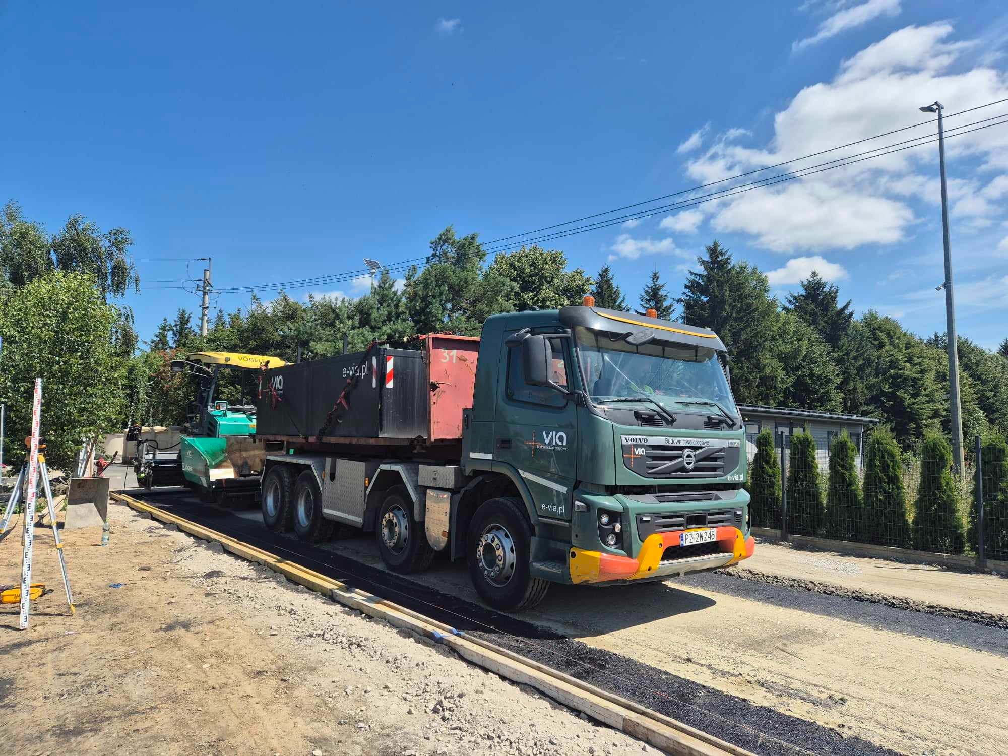 Zielona ciężarówka Volvo z logo VIA i adresem strony internetowej, obok rozściełacz asfaltu Vögele, na tle nieba i drzew, podczas prac drogowych.