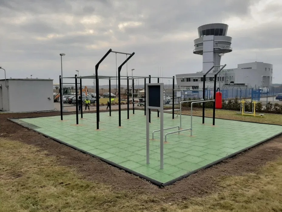 Zewnętrzna siłownia z zieloną nawierzchnią z gumowych płyt, widoczne stalowe konstrukcje do ćwiczeń, w tle budynek lotniska z wieżą kontroli lotów i pracownicy w odblaskowych kamizelkach.