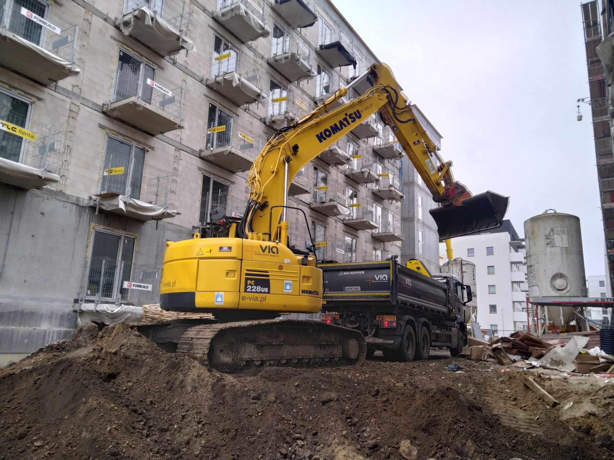 Żółta koparka Komatsu PC228US nabiera ziemię do czarnej wywrotki KH-Kipper na tle budowanego bloku z balkonami zabezpieczonymi siatką, widoczne oznaczenia 'FORBUILD' i 'TLC Rental'.