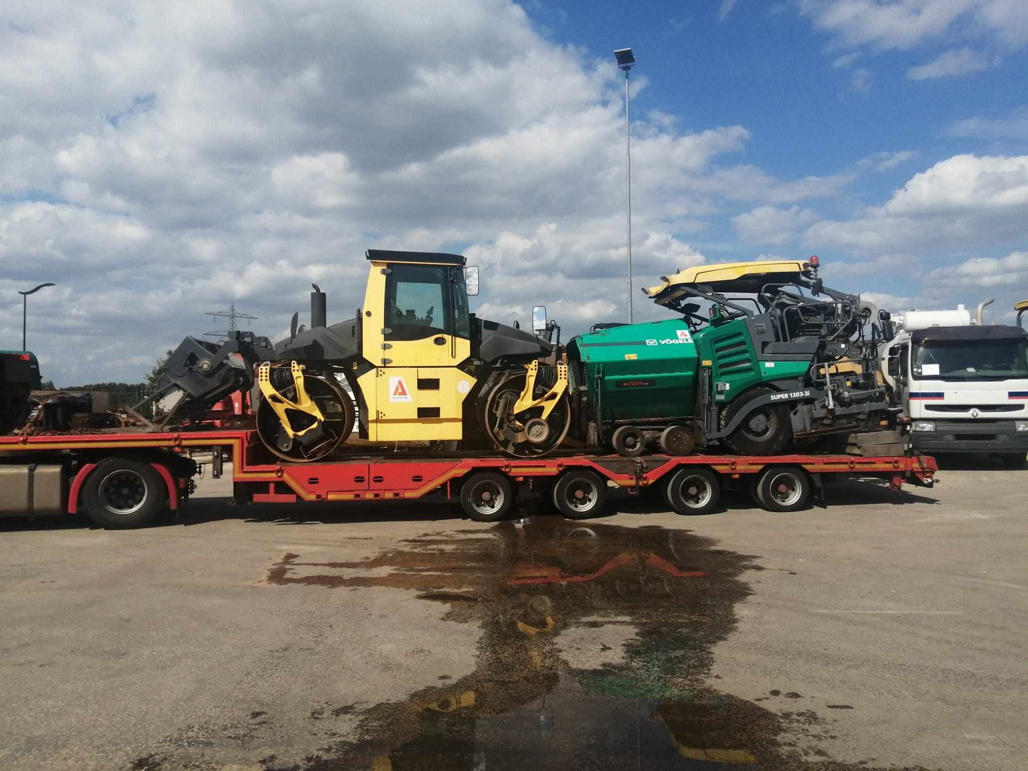 Żółty walec drogowy i zielony rozkładacz asfaltu marki Vogele Super 1303-3i przewożone na niskopodwoziowej platformie transportowej z czerwonymi elementami, stojącej na betonowym placu z kałużami