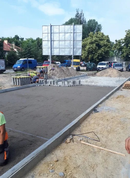 Przygotowanie podłoża pod nawierzchnię z kostki brukowej: wyrównany teren, obrzeża betonowe, pracownicy układający kostkę na obrzeżach, w tle zaparkowane samochody i tablica reklamowa.