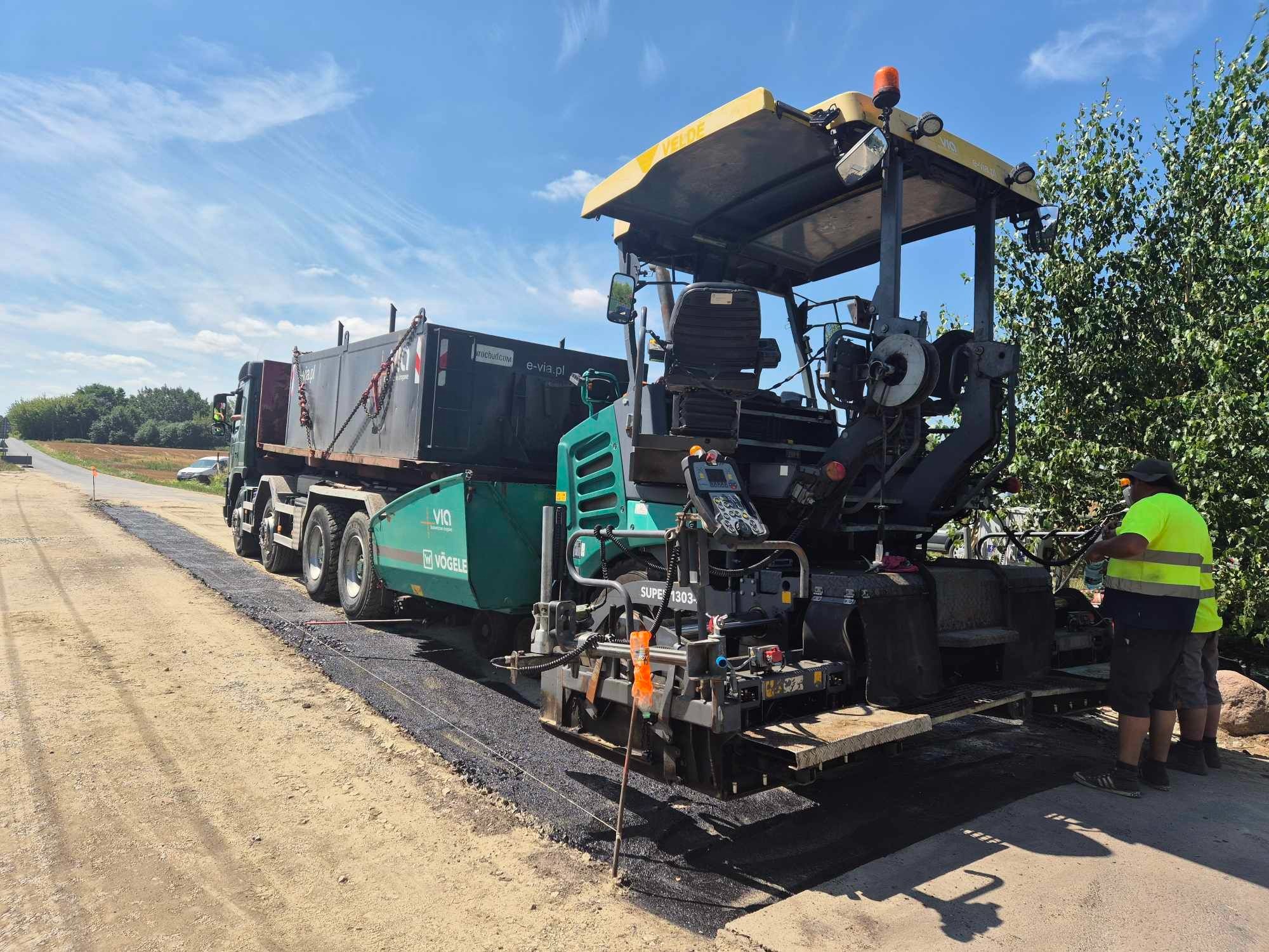 Rozściełacz asfaltu Vögele Super 1303-3 z podłączoną ciężarówką podczas układania nawierzchni asfaltowej na drodze gruntowej, widoczni dwaj pracownicy w odblaskowych kamizelkach.