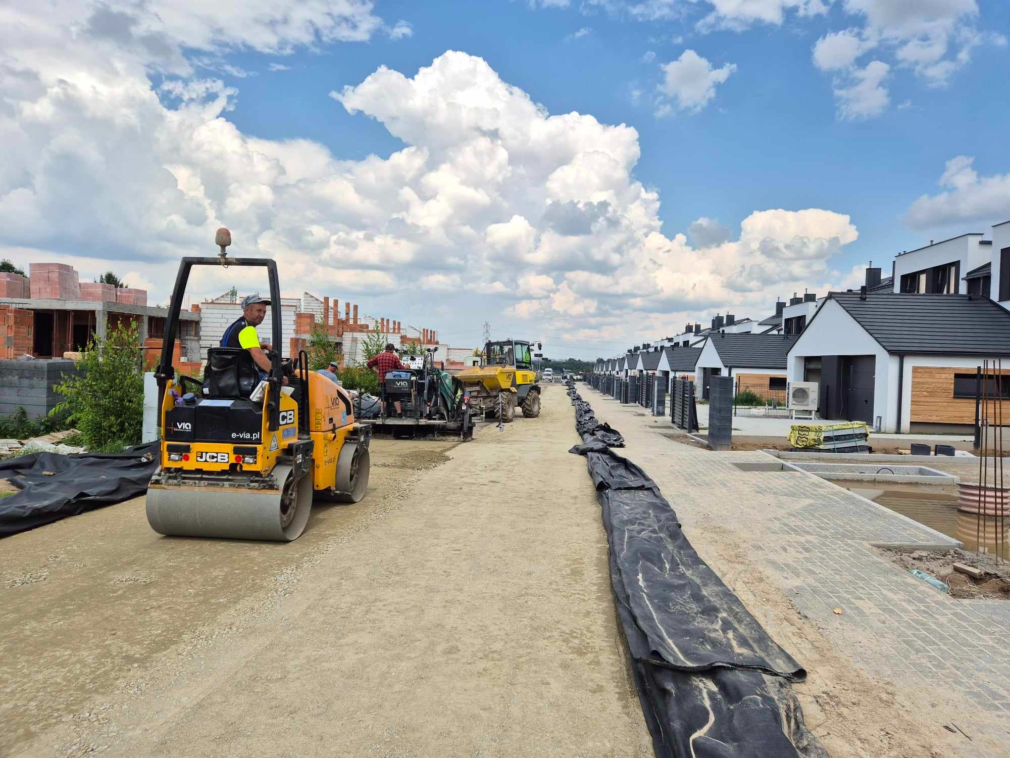Żółty walec drogowy JCB ubija nawierzchnię drogi gruntowej na tle rzędu nowych, szeregowych domów i błękitnego nieba z obłokami. W tle widoczne inne maszyny budowlane i robotnicy.